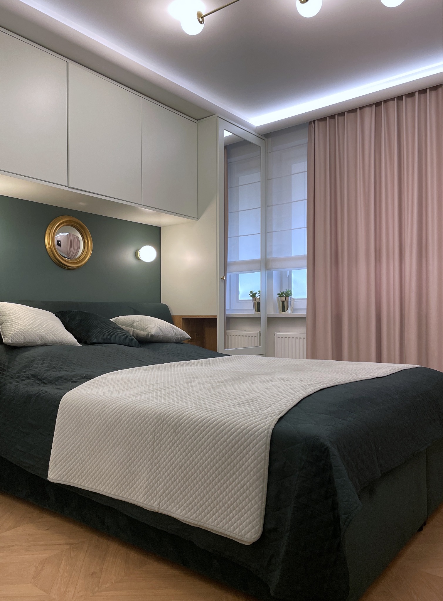 Nowoczesna sypialnia z łóżkiem w odcieniach zieleni, białymi szafkami nad łóżkiem i okrągłym lustrem w złotej ramie. Parkiet w jodełkę i pudrowo-różowe zasłony.