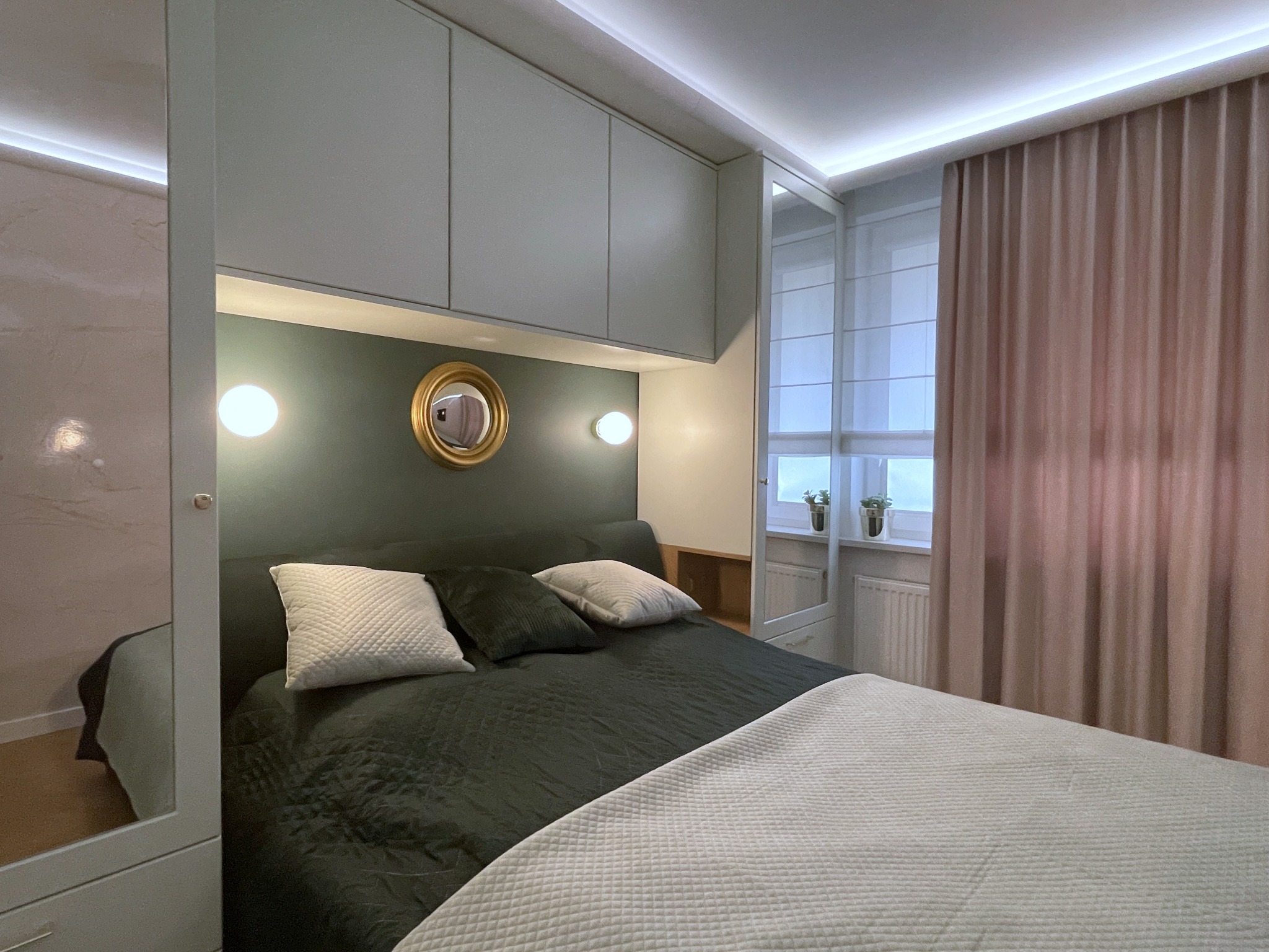 Nowoczesna sypialnia w stonowanych barwach: oliwkowa ściana za łóżkiem, okrągłe lustro w złotej ramie, białe szafki pod sufitem i pudrowo-różowe zasłony.