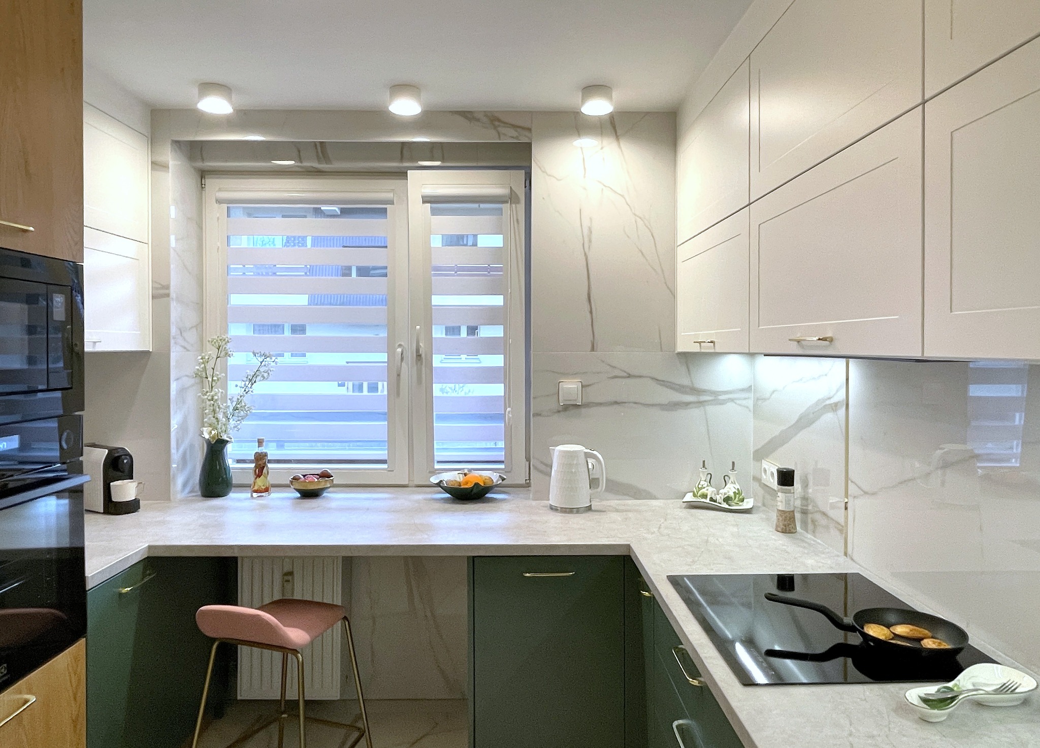 Nowoczesna kuchnia z zielonymi szafkami, blatem imitującym marmur i oknem z roletami. Na blacie akcesoria kuchenne i dekoracje. Na płycie grzewczej patelnia z jedzeniem.
