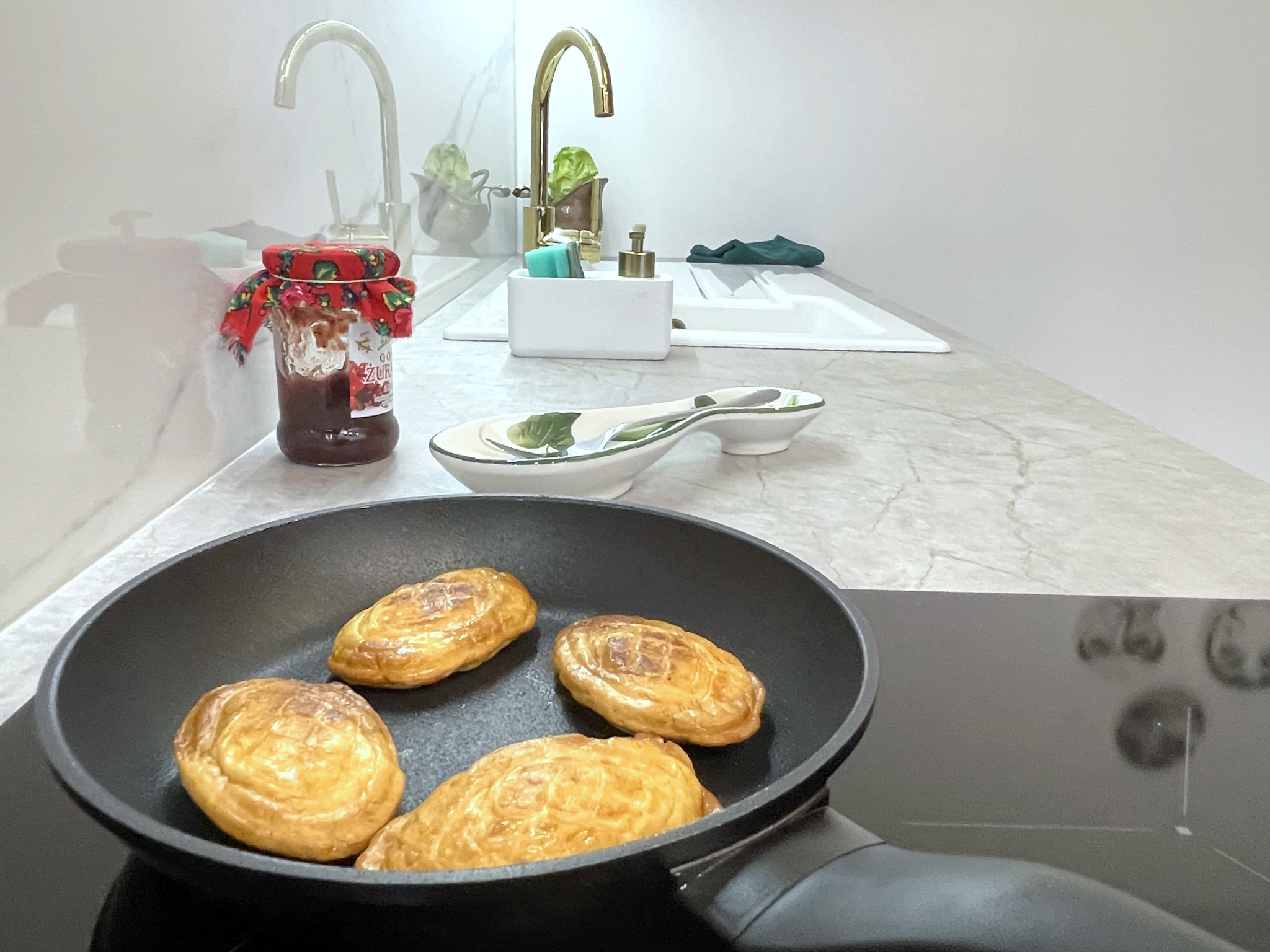 Nowoczesna kuchnia z marmurowym blatem: patelnia z pierogami na płycie indukcyjnej, złota bateria, słoik z dżemem w świątecznym ubranku i biały zlew.