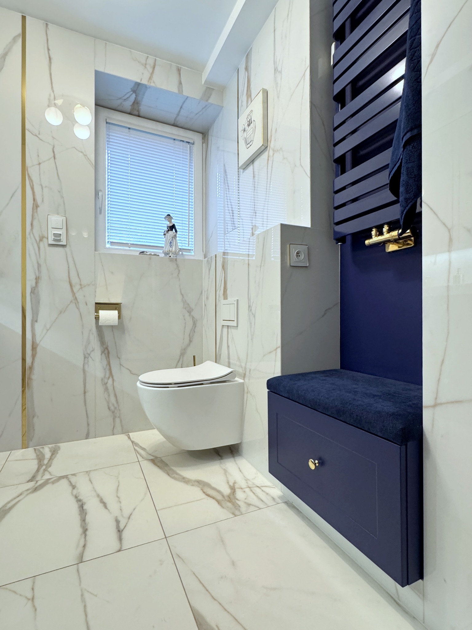 Elegancka łazienka w bieli i granacie: marmurowe płytki, wisząca toaleta, grzejnik drabinkowy i siedzisko z szufladą. Akcenty złota dodają luksusu.