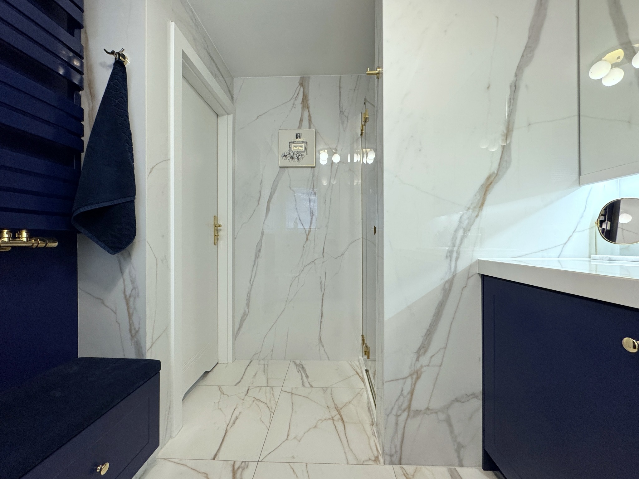 Elegancka łazienka z marmurowymi płytkami, granatowymi meblami i złotymi akcentami. Na ścianie dekoracja z motywem perfum. Widok na kabinę prysznicową.