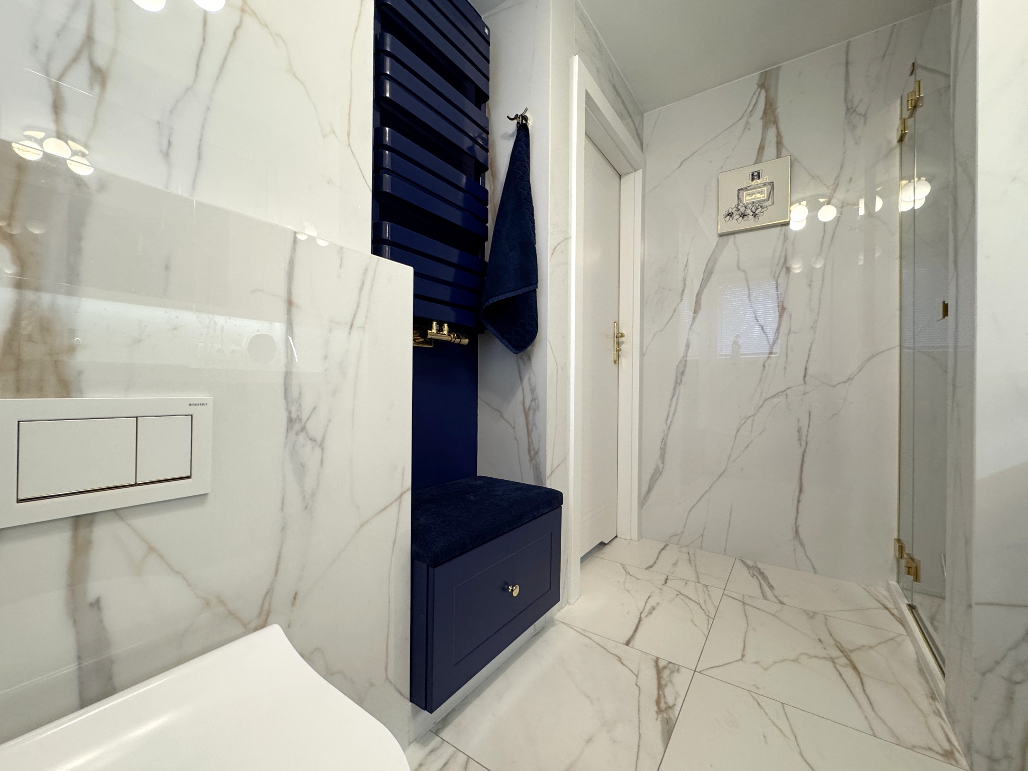 Nowoczesna łazienka z białymi płytkami imitującymi marmur, granatowym grzejnikiem drabinkowym i siedziskiem. Minimalistyczny design, złote akcenty.