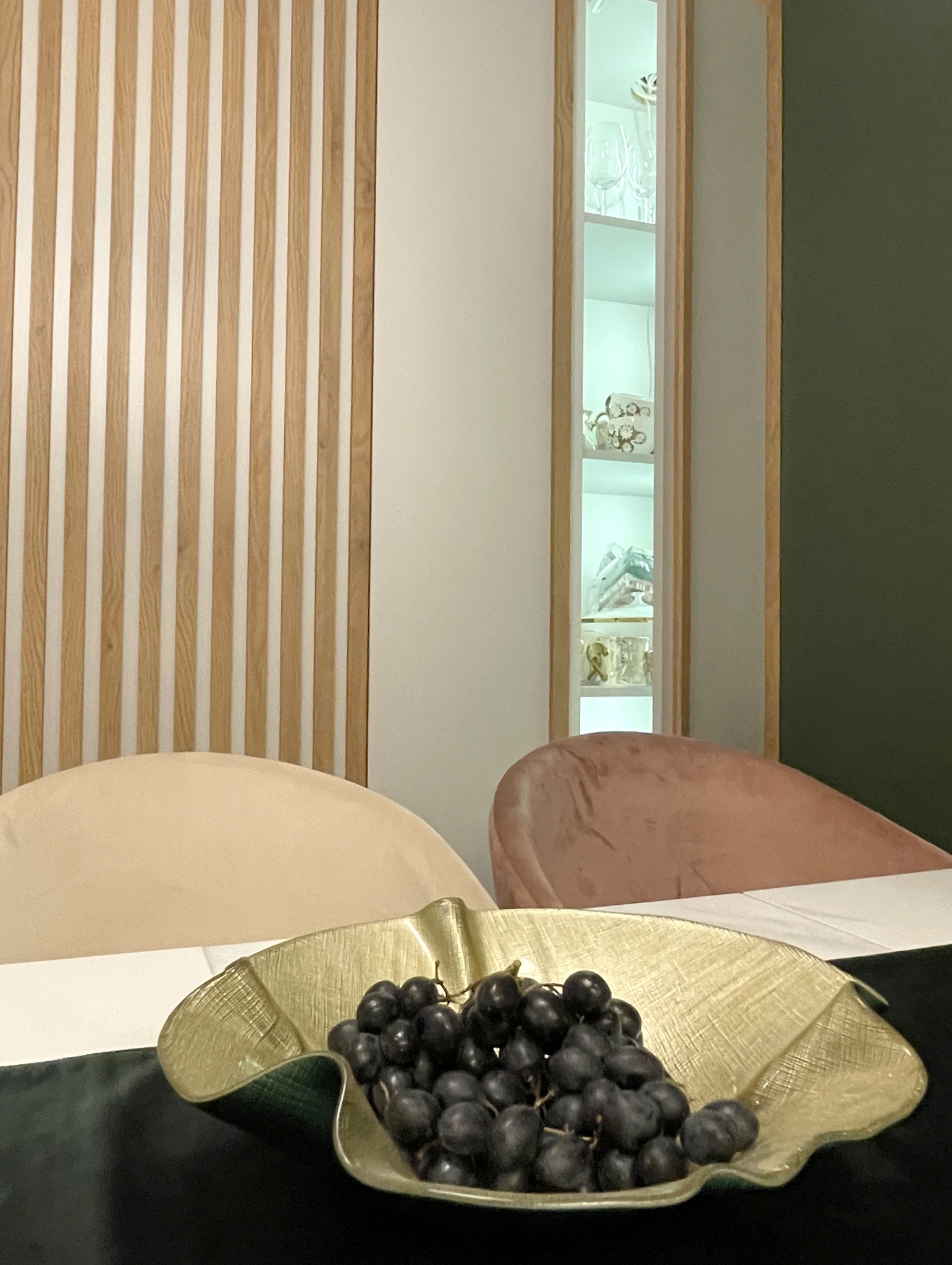 Stylowa aranżacja wnętrza z dekoracyjną ścianą z drewnianych listew, podświetlaną witryną i owocami w złotej misie na stole. Pastelowe krzesła w tle.