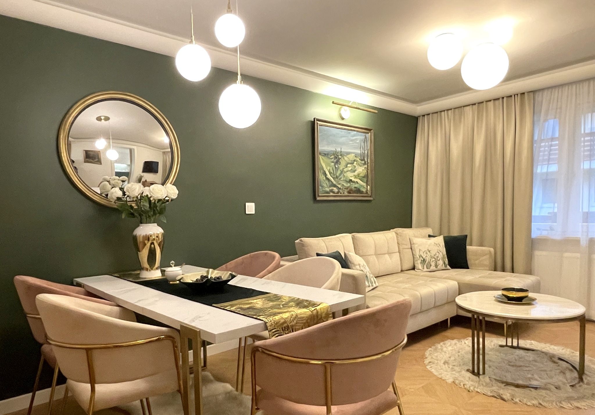 Elegancka aranżacja salonu z zieloną ścianą, okrągłym lustrem w złotej ramie, marmurowym stołem i beżową sofą. Nowoczesne lampy wiszące i obraz nadają wnętrzu charakteru.