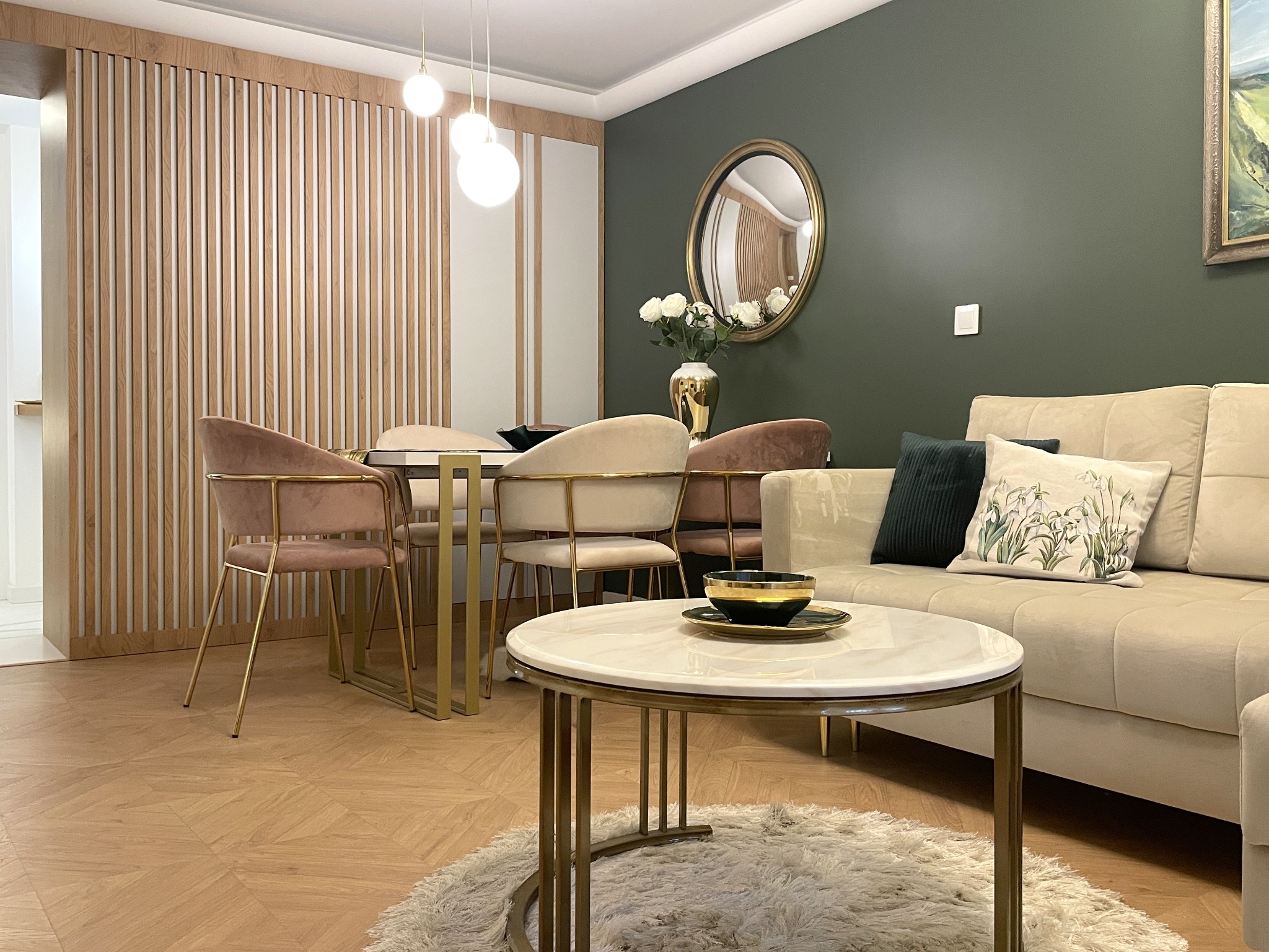 Salon w stylu glamour: beżowa sofa z poduszkami, okrągły stolik kawowy z marmurowym blatem, złote akcenty, drewniana ściana działowa i parkiet w jodełkę.