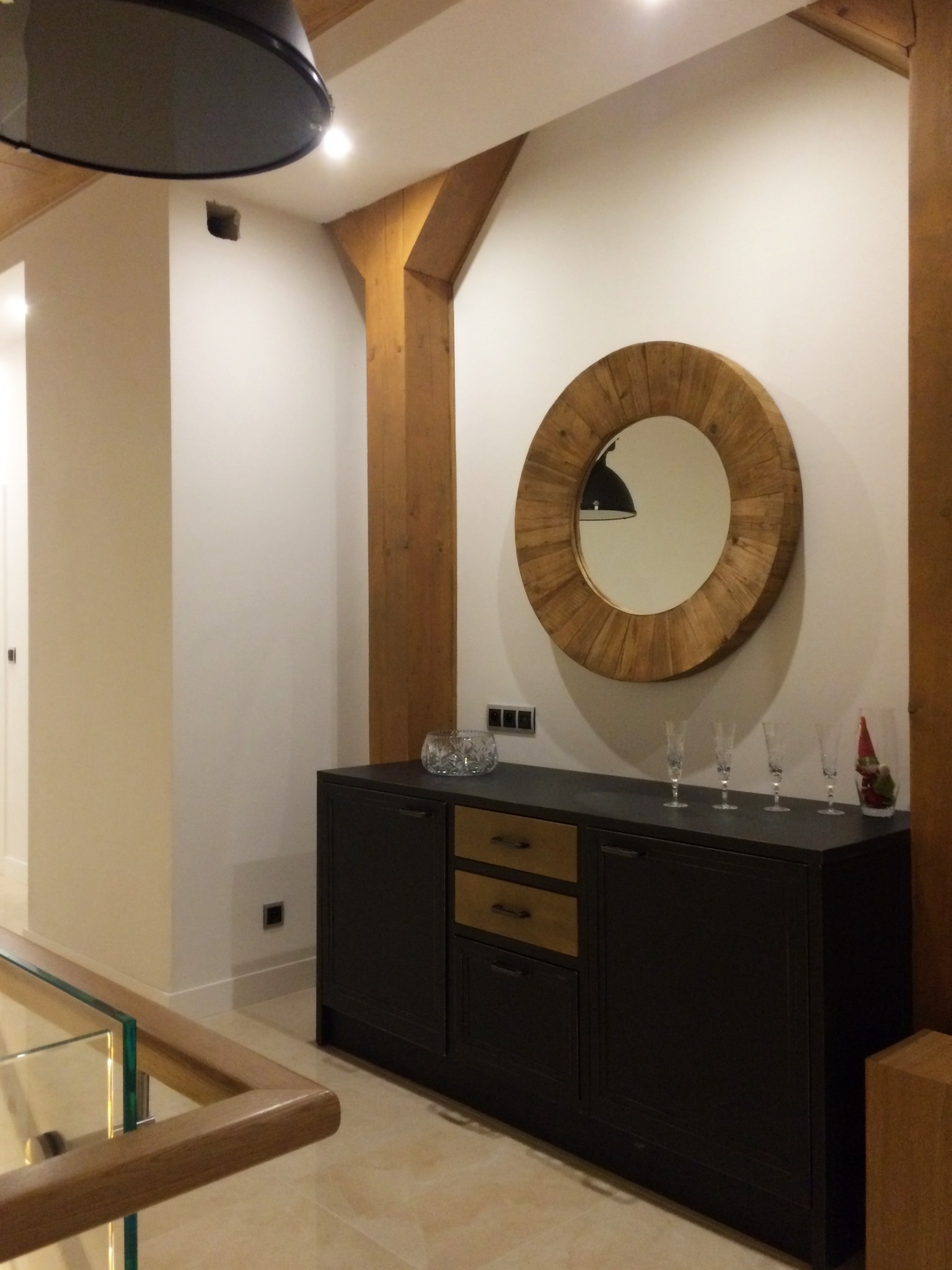 Hol na poddaszu z drewnianymi belkami stropowymi, okrągłym lustrem w drewnianej ramie nad czarną komodą z drewnianymi szufladami, na której stoją szklanki i ozdoba świąteczna.