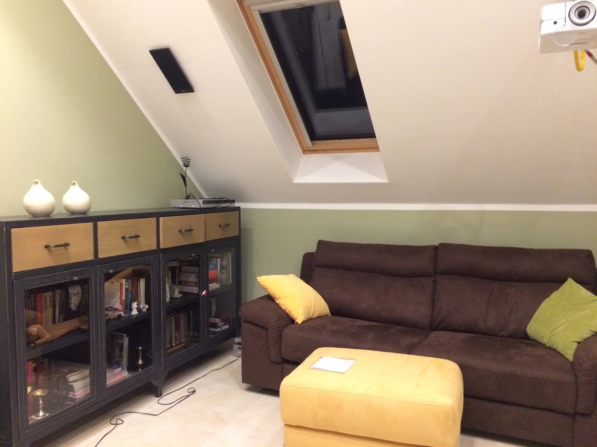 Pokój na poddaszu z projektorem, brązową kanapą, żółtym pufem i regałem z książkami, oświetlony światłem z okna dachowego.