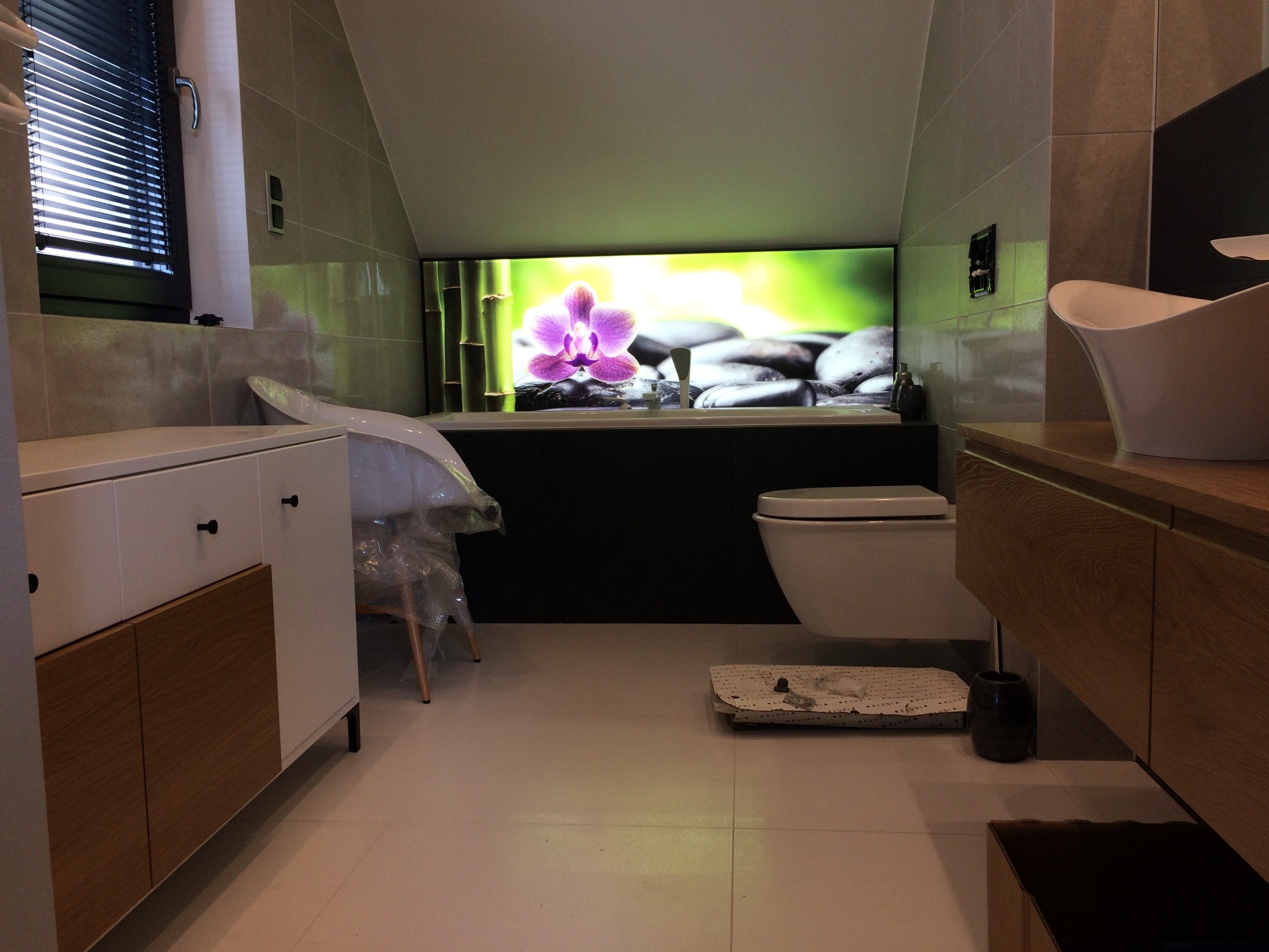 Łazienka na poddaszu z oknem nad wanną z motywem orchidei i kamieni zen, biała umywalka nablatowa, drewniane szafki i białe meble łazienkowe z czarnymi uchwytami.