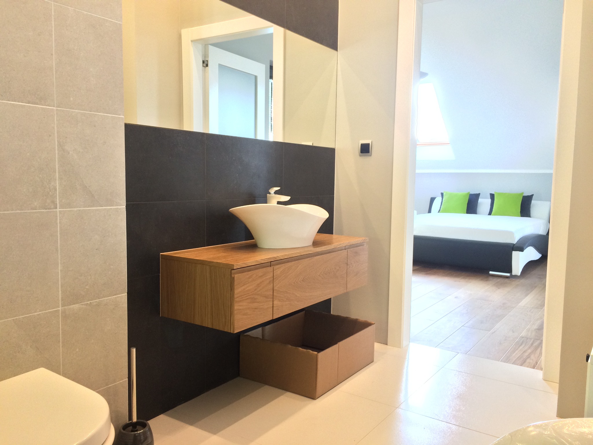 Nowoczesna łazienka z umywalką nablatową w kształcie misy, drewniana szafka pod umywalką, czarne płytki za umywalką, widok na sypialnię z poddaszem w tle.