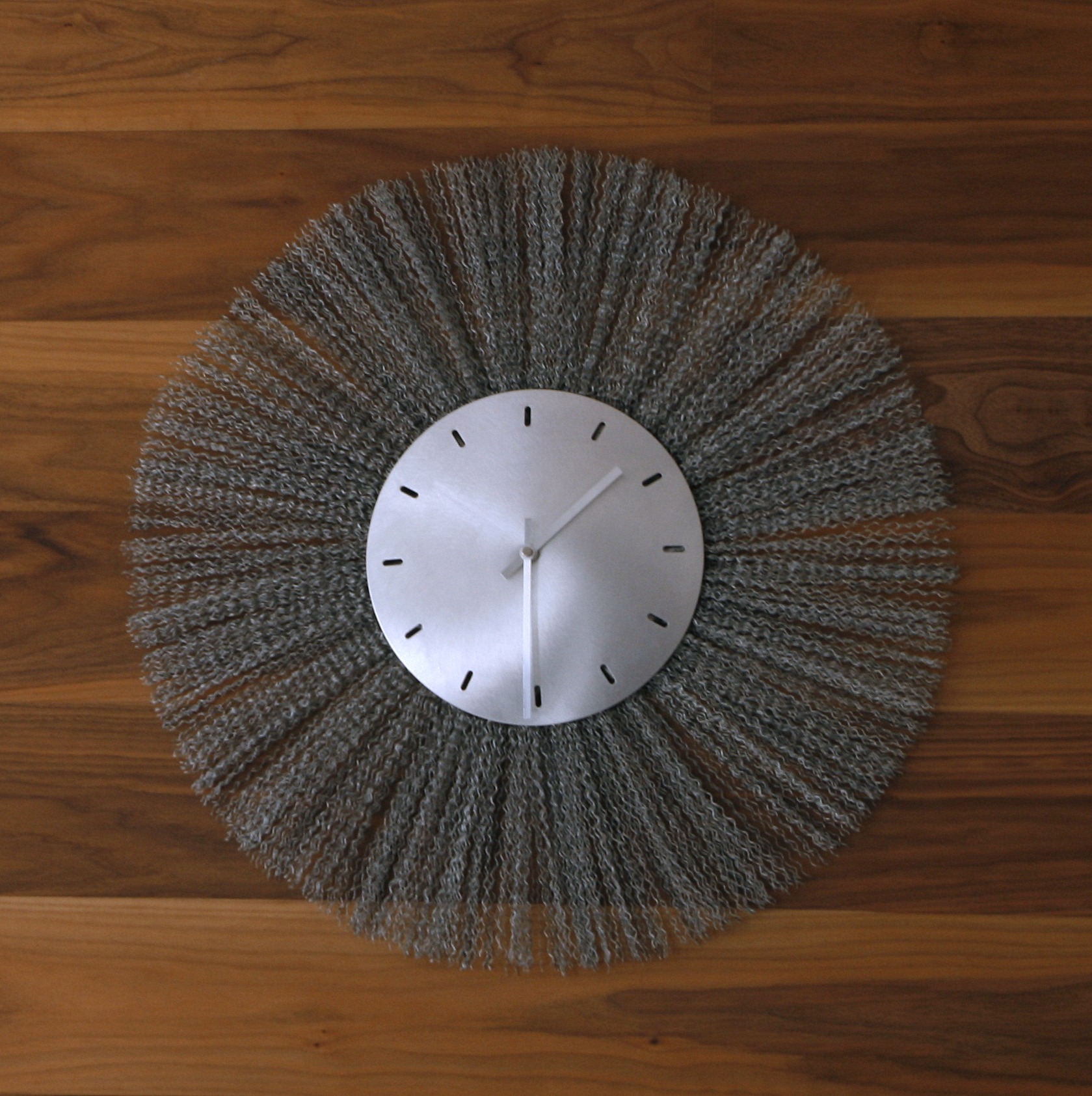 Nowoczesny zegar ścienny z metalową tarczą i dekoracyjnym, szarym, włóczkowym obramowaniem na tle drewnianej ściany.