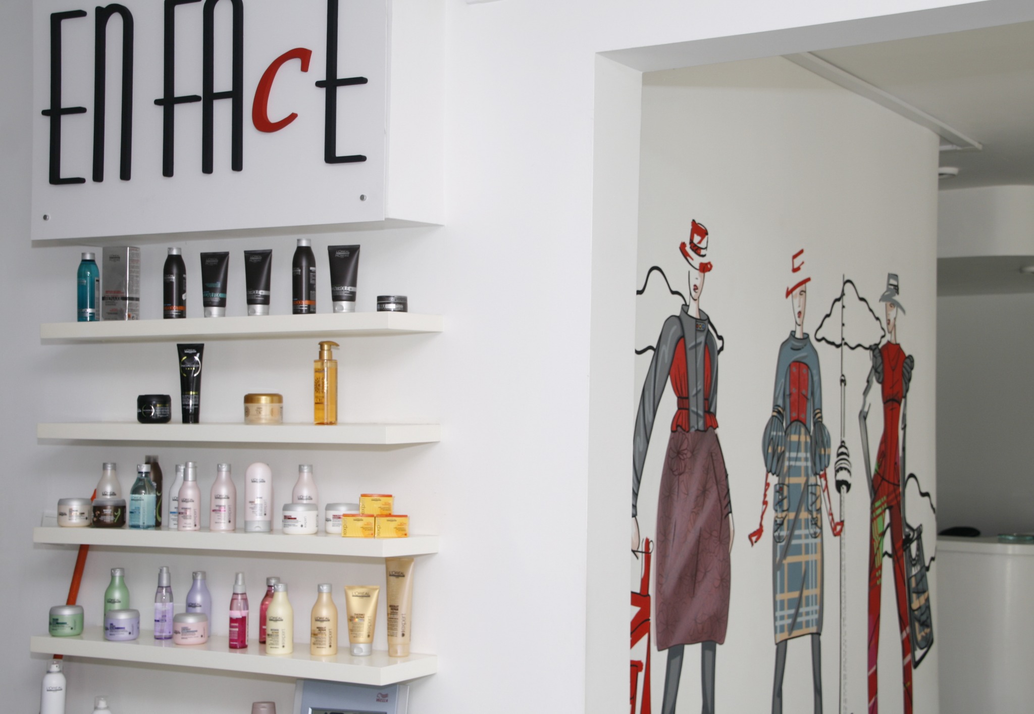 Wnętrze salonu fryzjerskiego: białe ściany, półki z kosmetykami do włosów marki L'Oreal, grafika przedstawiająca modelki w kapeluszach i sukienkach.