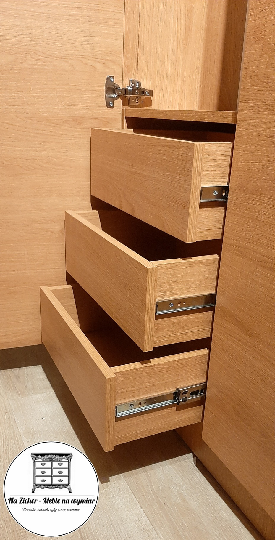 Wnętrze szafy z trzema wysuniętymi szufladami z jasnego drewna, widoczne metalowe prowadnice, zawias drzwi szafy w górnym rogu, na dole w rogu okrągłe logo firmy.