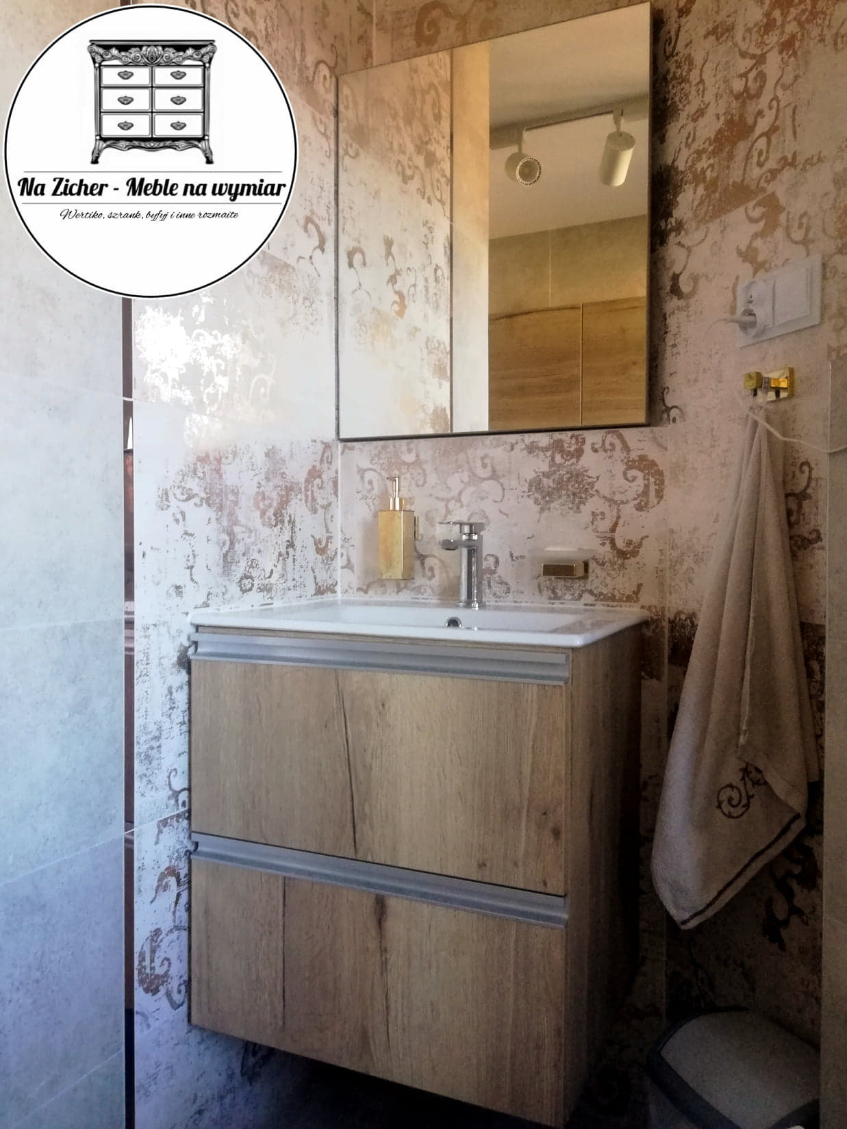 Szafka łazienkowa z umywalką w kolorze drewna, z dwoma szufladami, wisząca na tle ściany z wzorzystymi płytkami, obok lustro z szafką i ręcznik z haftowanym wzorem.
