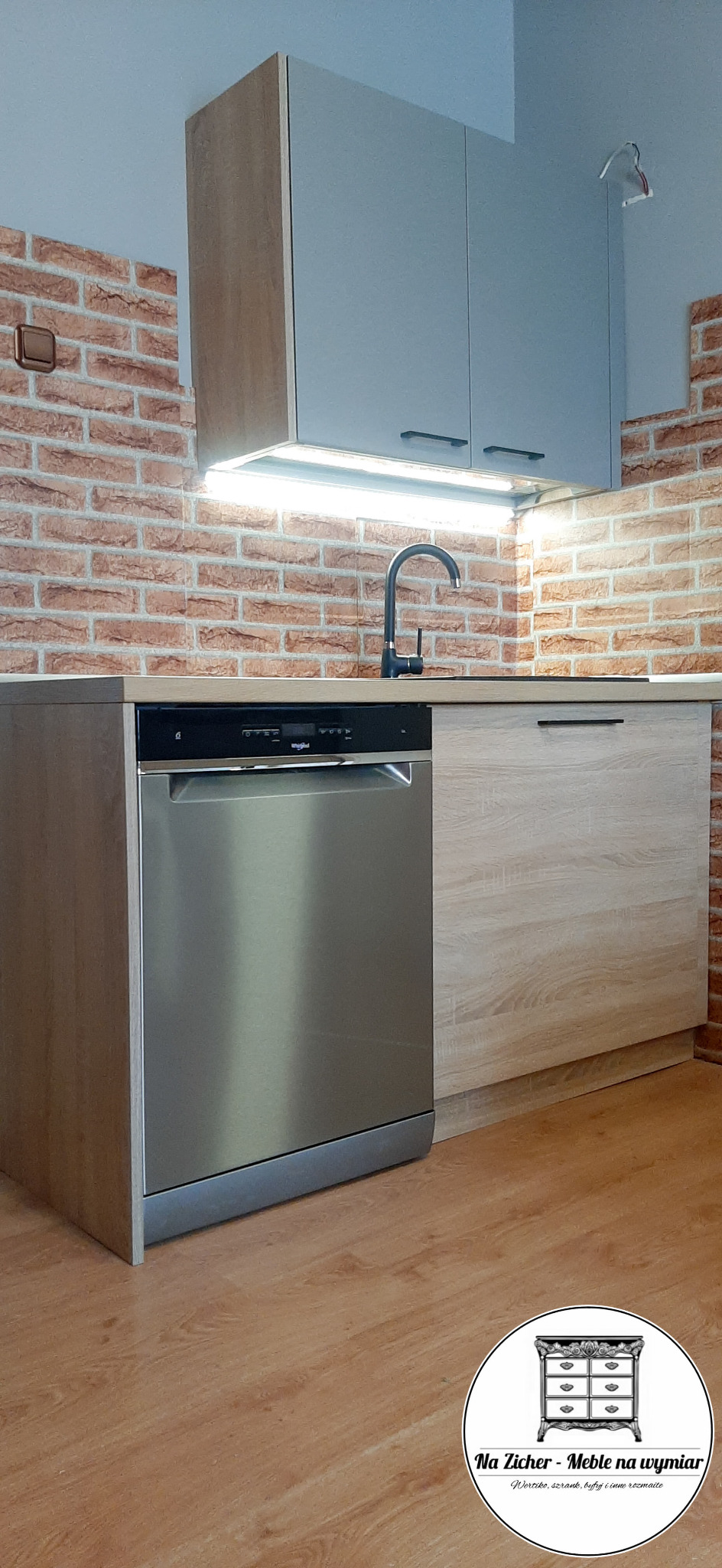 Fragment kuchni z szarymi szafkami wiszącymi, blatem w odcieniu jasnego drewna i srebrną zmywarką; ściana wyłożona ceglastymi płytkami, czarna bateria kuchenna