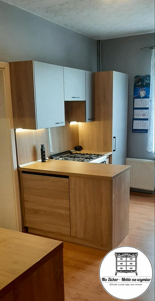 Fragment szarej kuchni z drewnianymi akcentami, oświetlenie LED pod szafkami, widoczna płyta gazowa i logo firmy w rogu kadru.