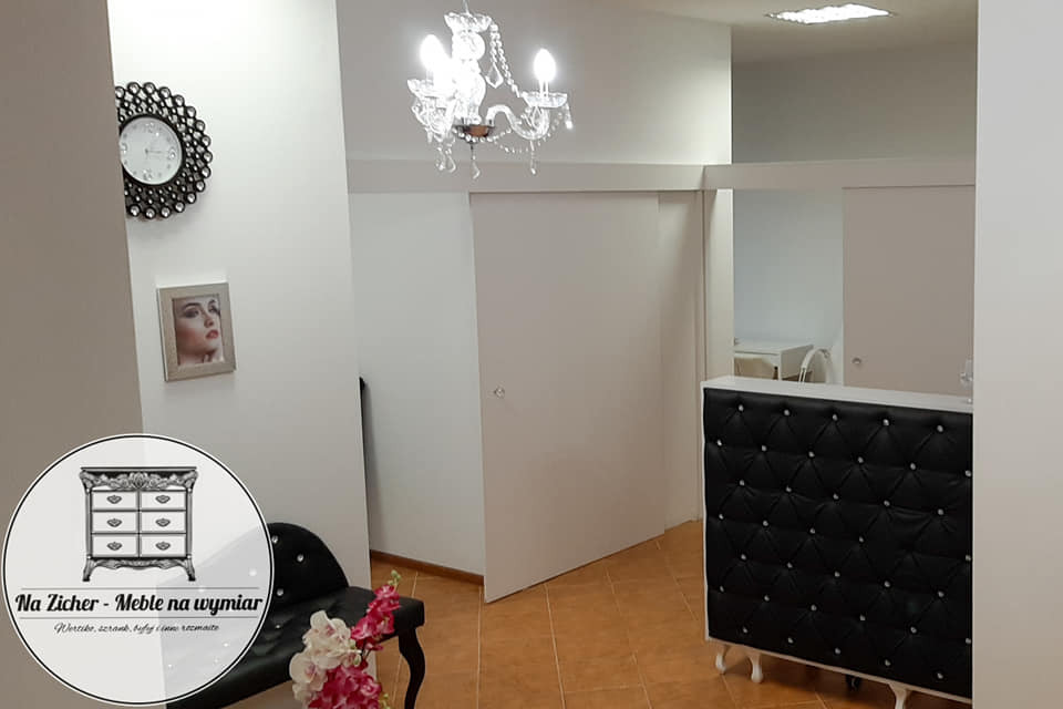 Wnętrze z przesuwnymi białymi drzwiami, kryształowym żyrandolem, czarnym fotelem z pikowanym oparciem i logo firmy meblarskiej w rogu.