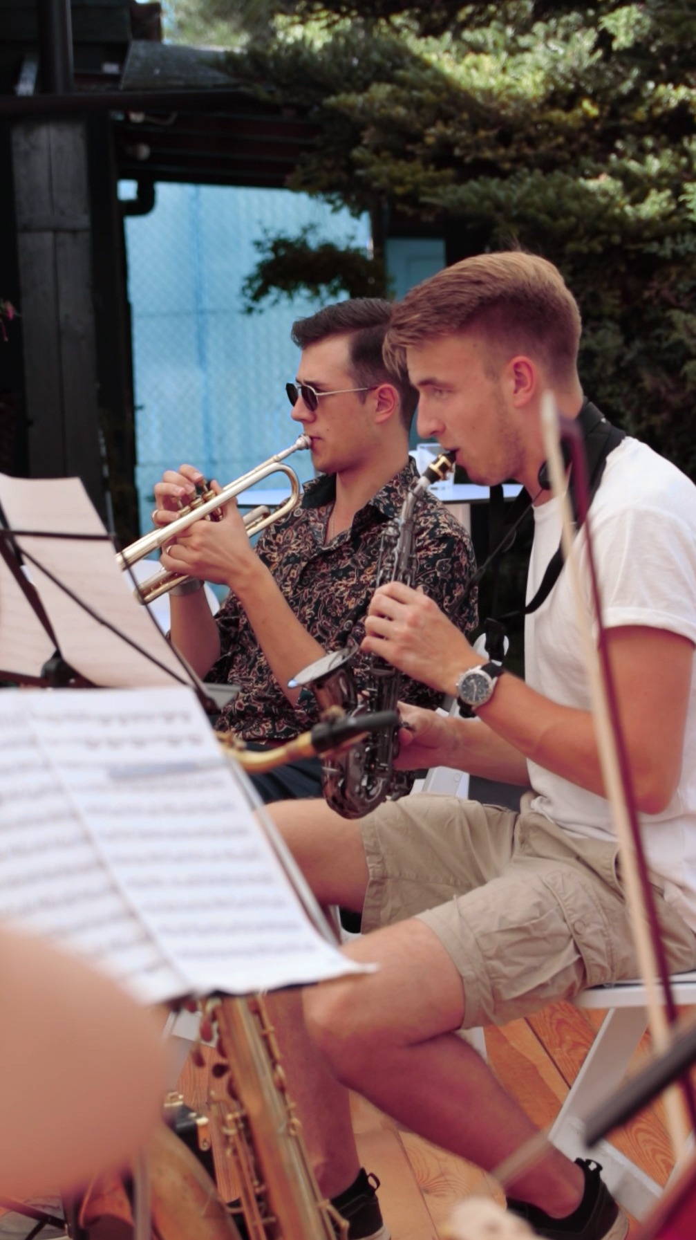 Dwóch młodych mężczyzn gra na instrumentach dętych podczas koncertu plenerowego: jeden na trąbce, drugi na saksofonie. Obaj siedzą na krzesłach, przed nimi nuty na statywach.