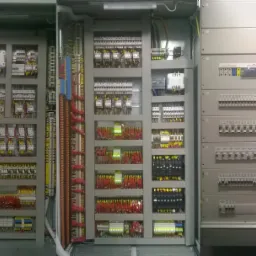 Rozbudowana, otwarta szafa sterownicza z licznymi przewodami, bezpiecznikami i elementami elektrycznymi ułożonymi w regularny sposób.