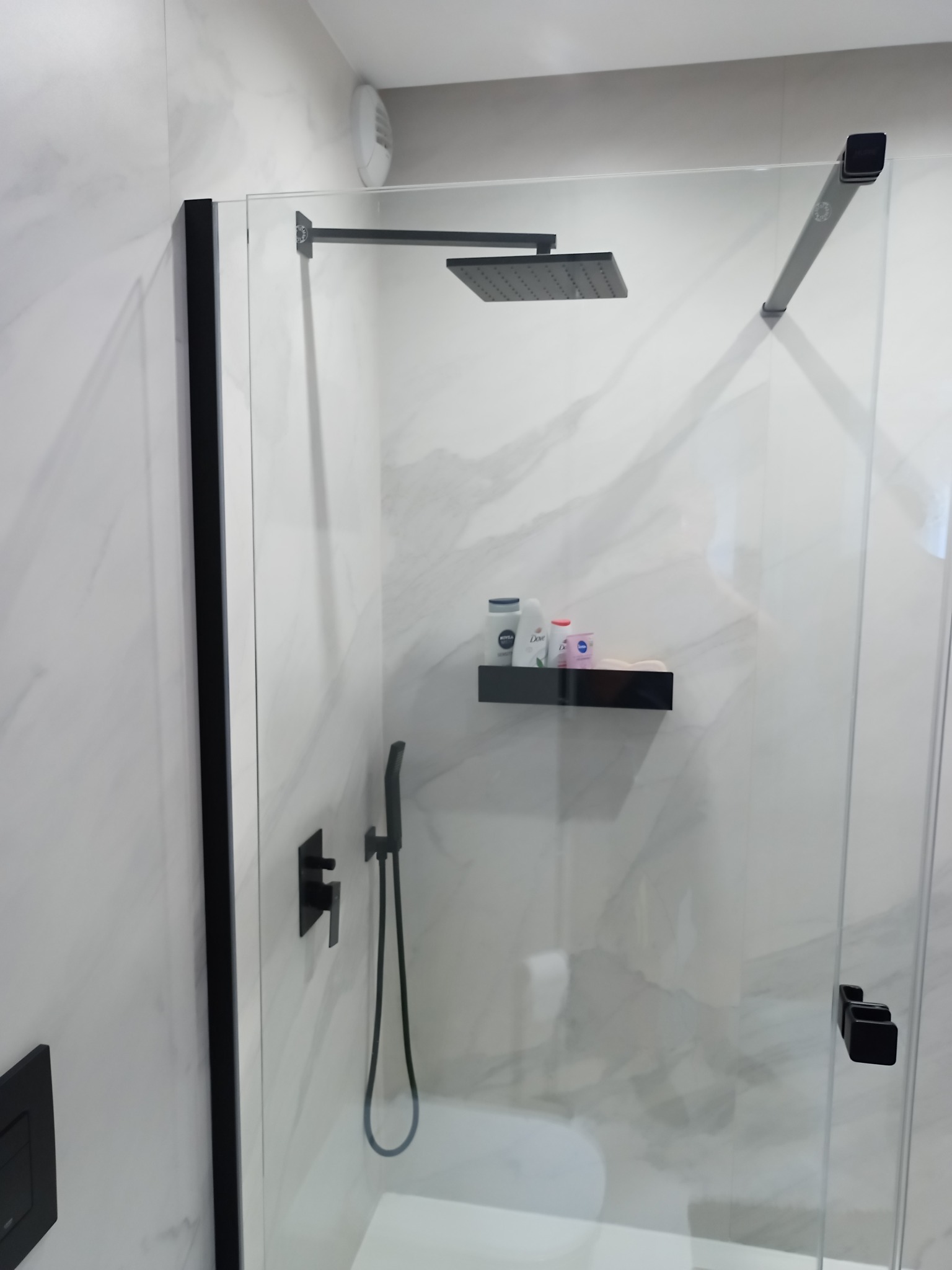 Nowoczesna kabina prysznicowa walk-in z czarnymi elementami, marmurowe płytki na ścianach, półka z kosmetykami. Minimalistyczny design, eleganckie wykończenie.