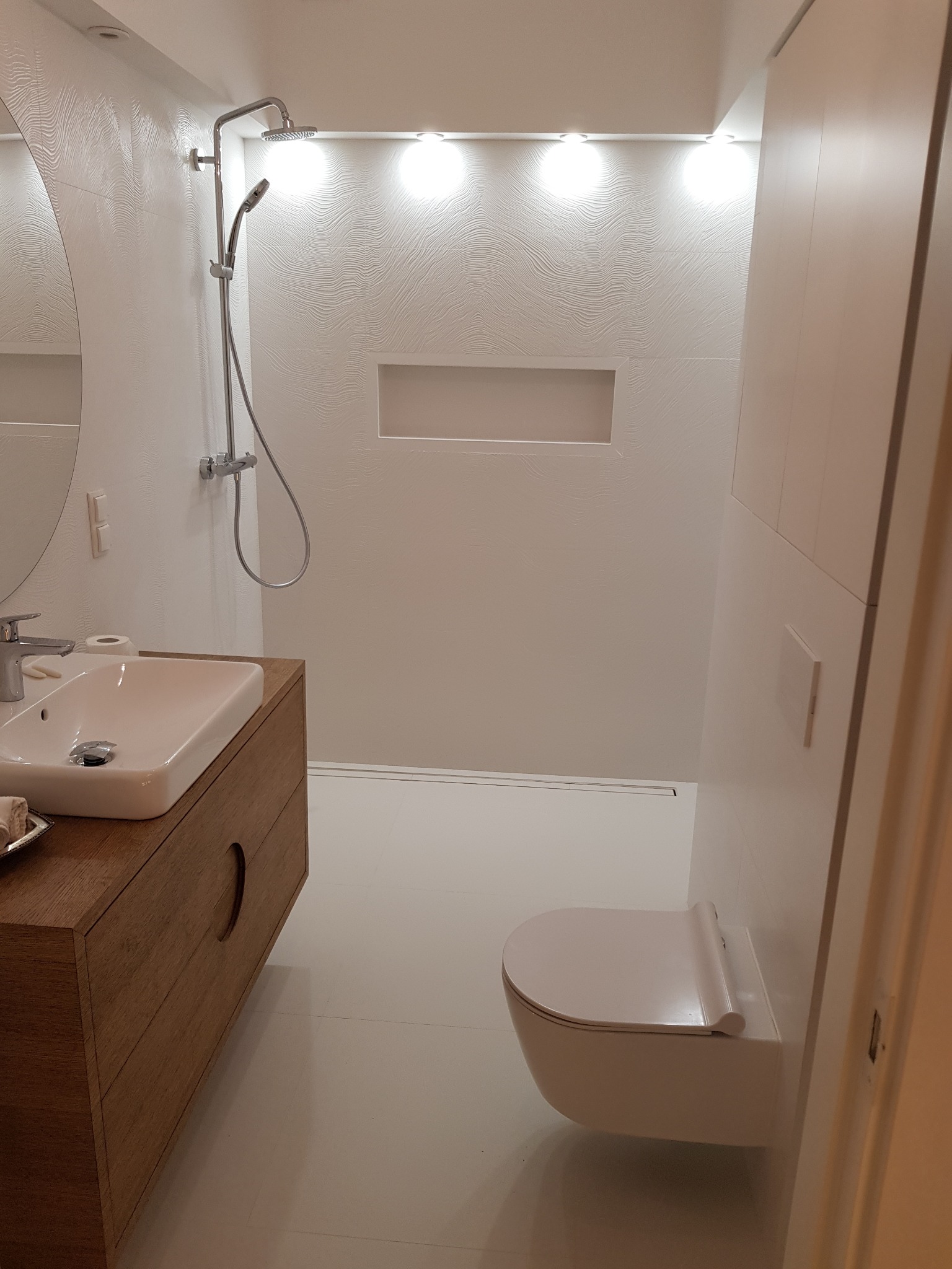 Nowoczesna łazienka z białymi płytkami o falistej strukturze, drewnianą szafką pod umywalką i podwieszaną toaletą, oświetlona punktowymi lampami sufitowymi.