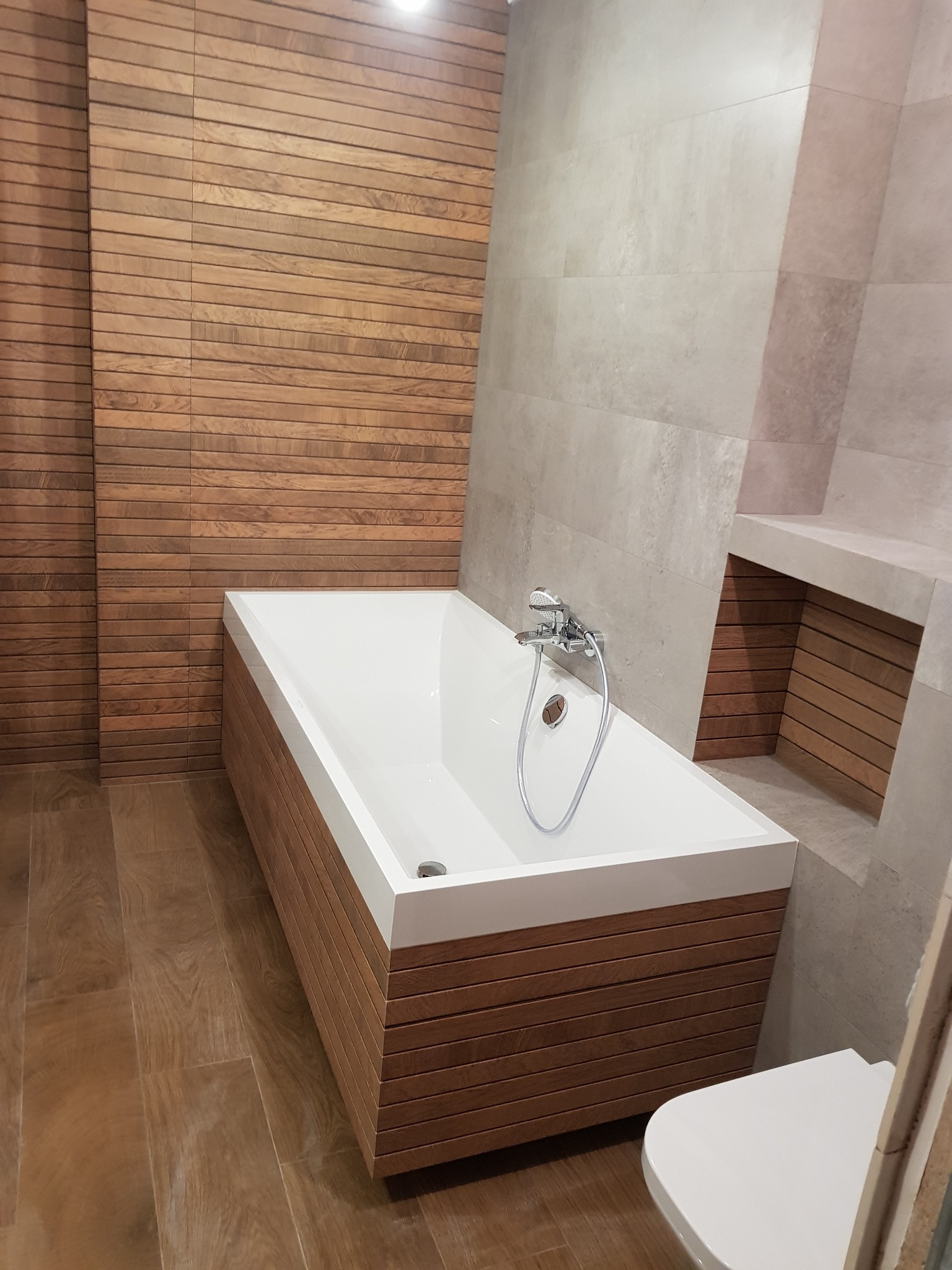 Nowoczesna łazienka z wanną obudowaną drewnopodobnymi panelami, szare płytki na ścianach i drewnopodobna podłoga, minimalistyczna armatura.