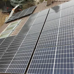 Brązowy dach z zamontowanymi panelami słonecznymi, widoczne okna dachowe i drabina oparta o jeden z paneli.