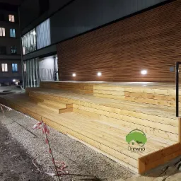 Drewniany taras z podestem i schodami przed budynkiem, oświetlony punktowymi lampami, otoczony szarym żwirem i taśmą ostrzegawczą, z widocznym logo firmy na pierwszym planie.