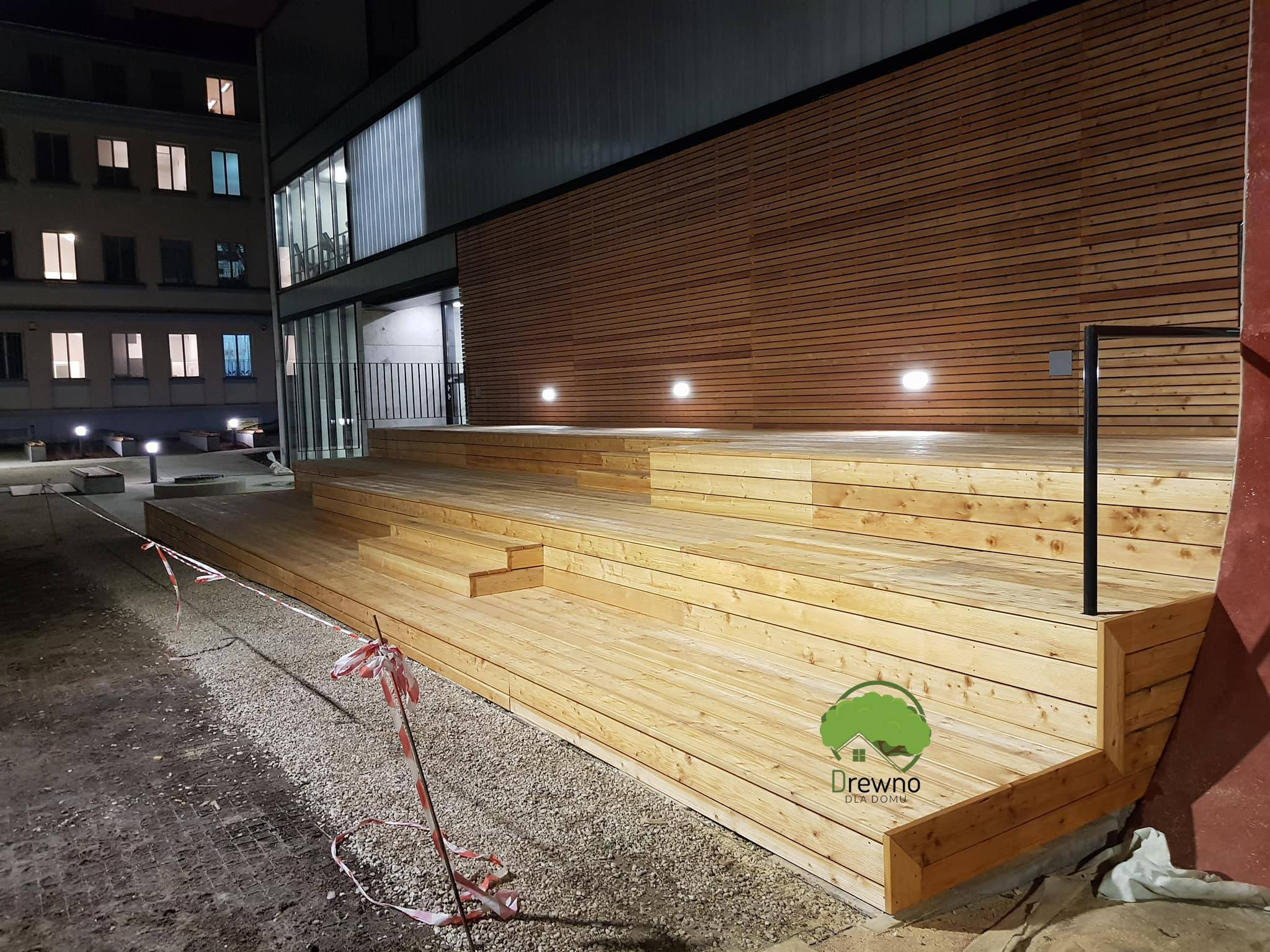 Drewniany taras z podestem i schodami przed budynkiem, oświetlony punktowymi lampami, otoczony szarym żwirem i taśmą ostrzegawczą, z widocznym logo firmy na pierwszym planie.
