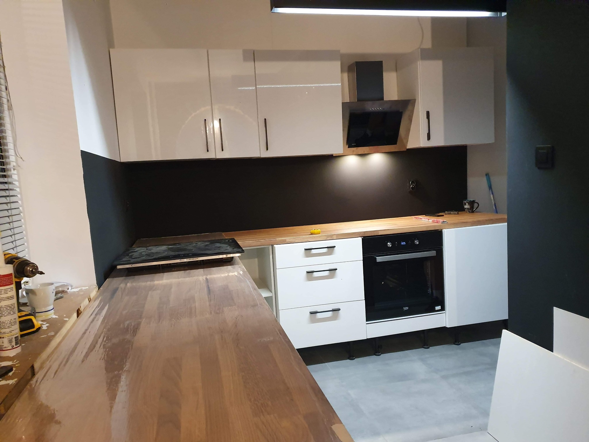 Nowoczesna kuchnia w trakcie montażu: białe szafki z czarnymi uchwytami, drewniany blat roboczy z widocznym klejem i narzędziami, czarny panel nad blatem, płyta indukcyjna i piekarnik w zabudowie.