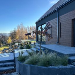 Klim-Bruk Piotr Klimca - Nowoczesny dom z tarasem wyłożonym szarą kostką, ozdobiony trawami ozdobnymi i drzewkiem. Widok na okolicę w słoneczny dzień.