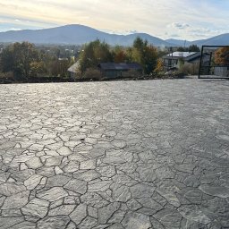 Klim-Bruk Piotr Klimca - Ukończona powierzchnia z szarej kostki brukowej o nieregularnym kształcie na tle górskiego krajobrazu i częściowo zamontowanej, czarnej bramy wjazdowej.