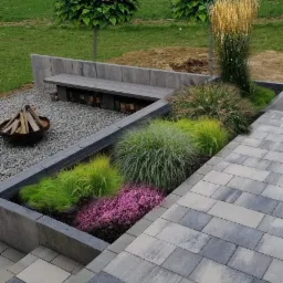 Ułożona z szarych kostek ścieżka prowadzi do betonowej rabaty z różnokolorową roślinnością, obok palenisko z drewnem i ławka z zapasem drewna w tle.