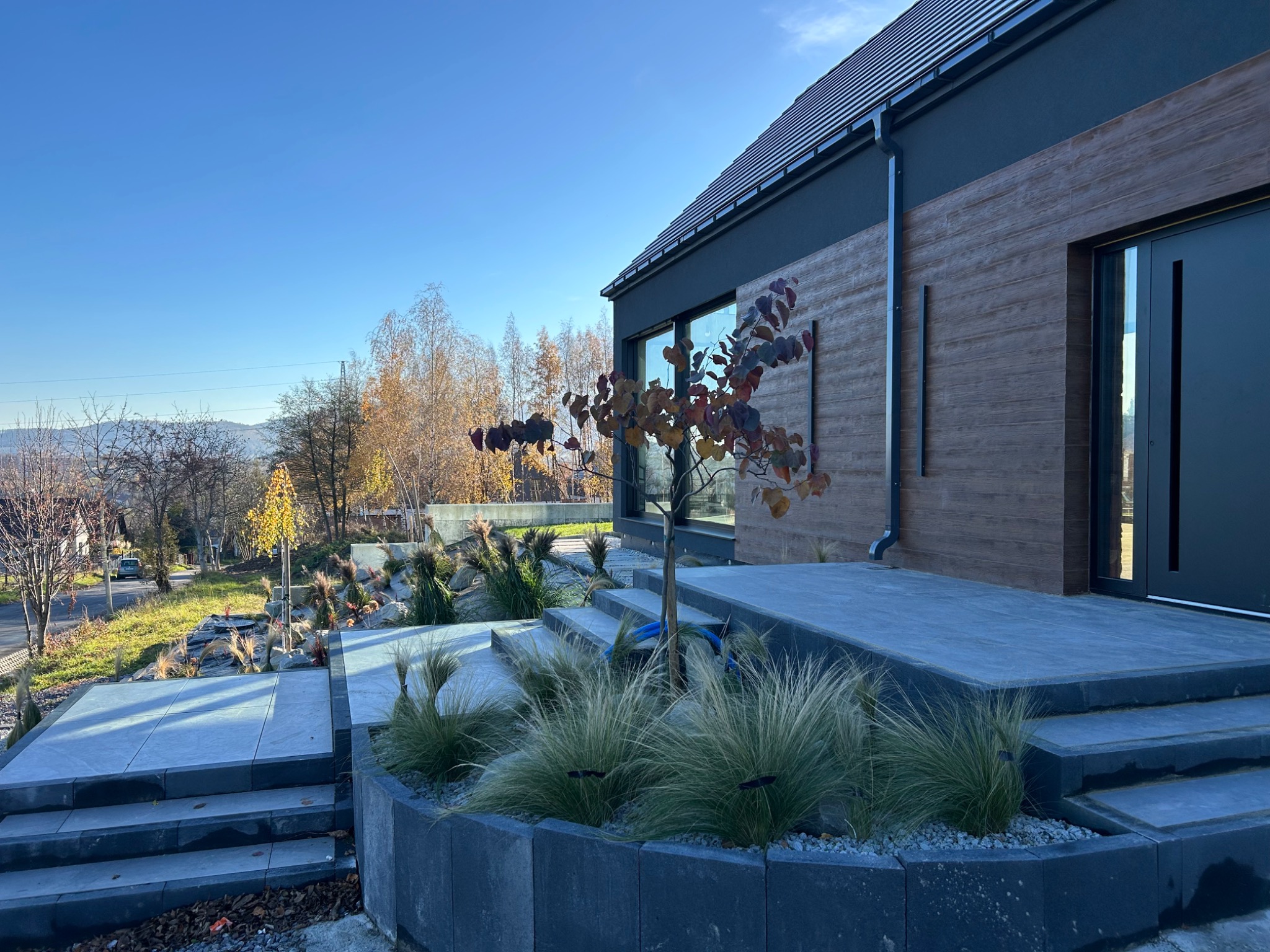 Nowoczesny dom z tarasem wyłożonym szarą kostką, ozdobiony trawami ozdobnymi i drzewkiem. Widok na okolicę w słoneczny dzień.