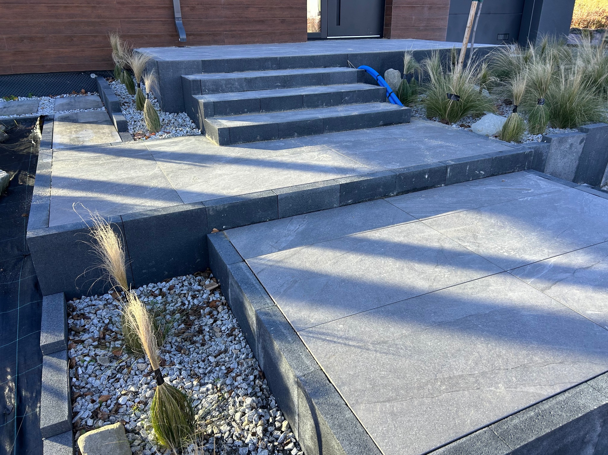 Nowoczesne schody wejściowe i taras z szarych płyt, obrzeża z kostki. Dekoracyjne trawy i kamień ozdobny. Niebieski przewód na schodach.
