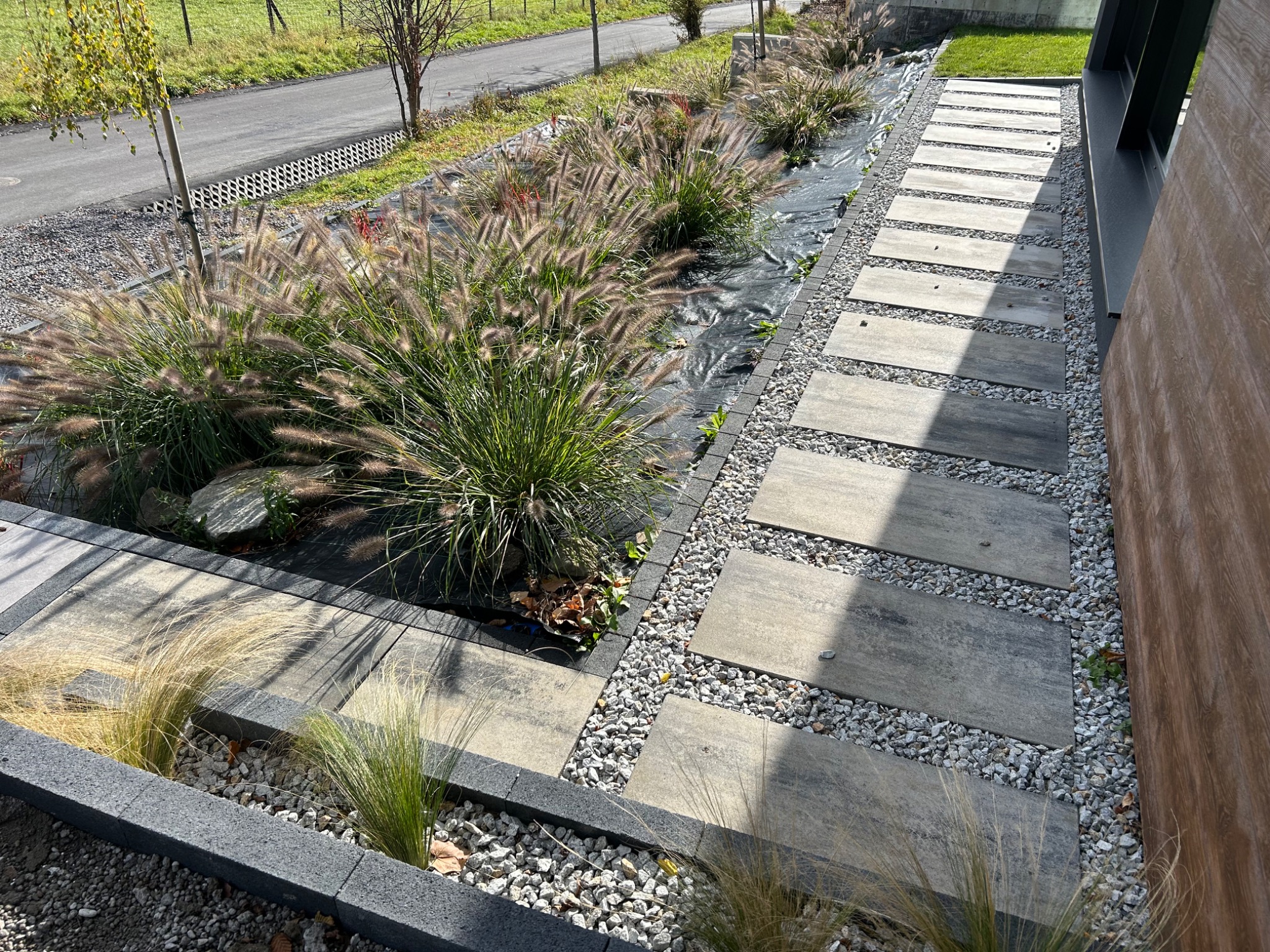Ścieżka z płyt betonowych oddzielona żwirem, prowadząca przez ogród z ozdobnymi trawami. Czarna agrowłóknina widoczna pod roślinami. Krawężniki z szarego betonu.