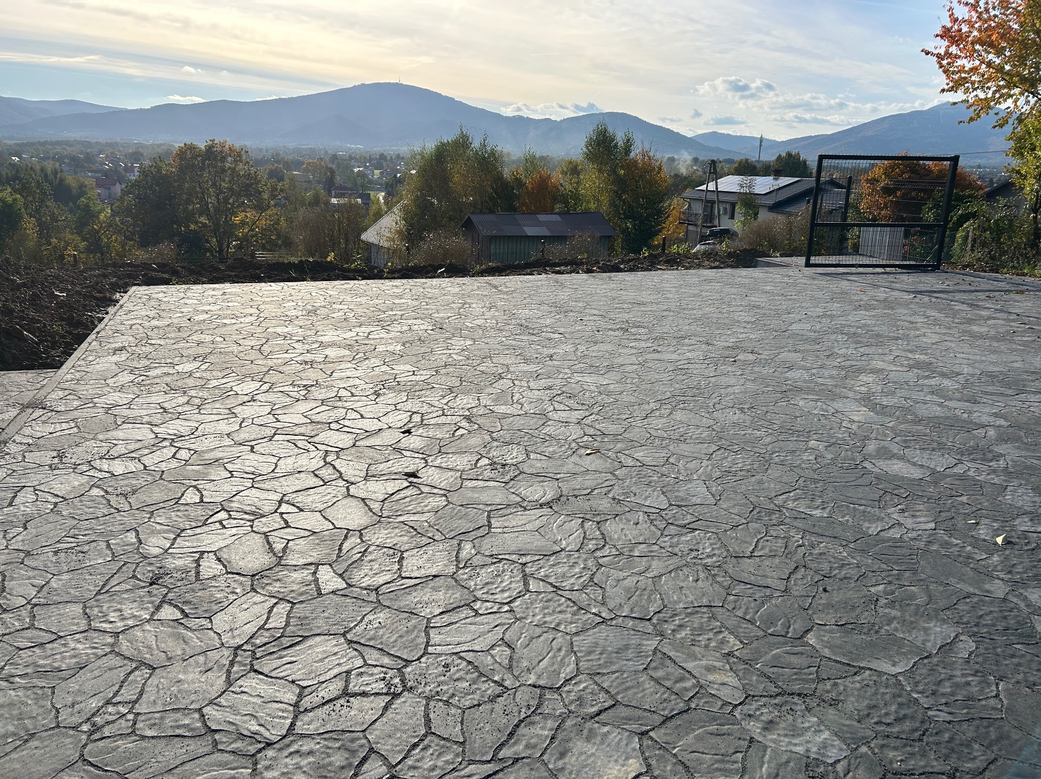 Ukończona powierzchnia z szarej kostki brukowej o nieregularnym kształcie na tle górskiego krajobrazu i częściowo zamontowanej, czarnej bramy wjazdowej.