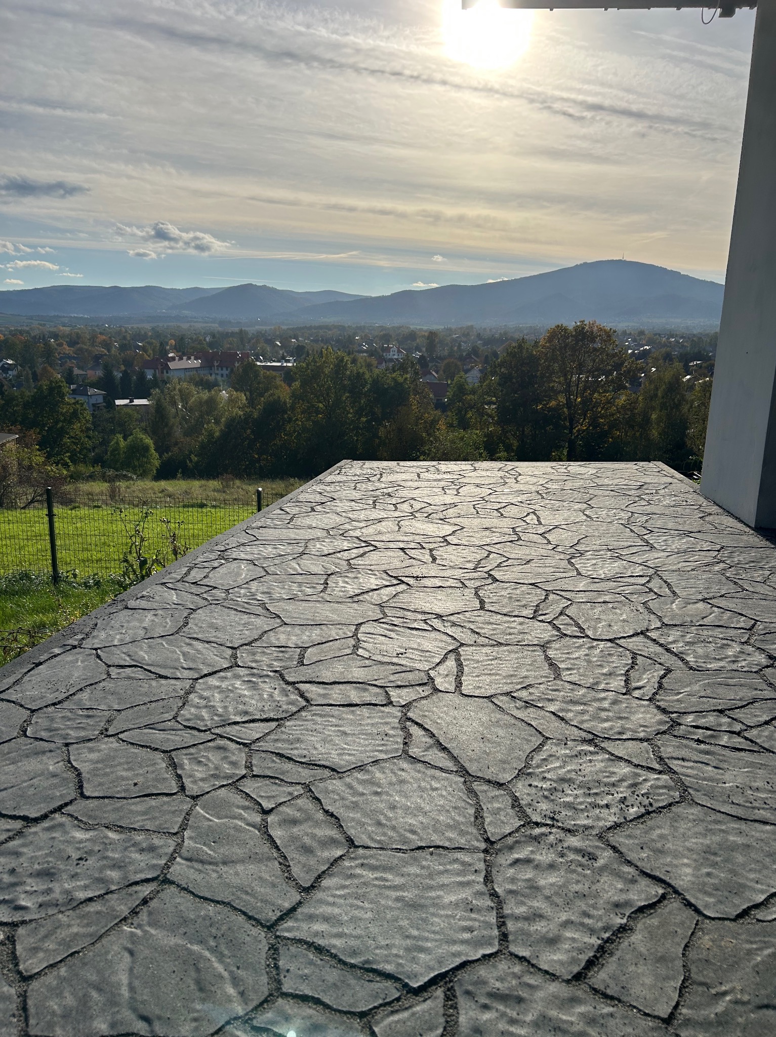 Szara, nieregularna nawierzchnia z kamienia na tle krajobrazu z górami i zabudowaniami. Widok z góry, słoneczny dzień.