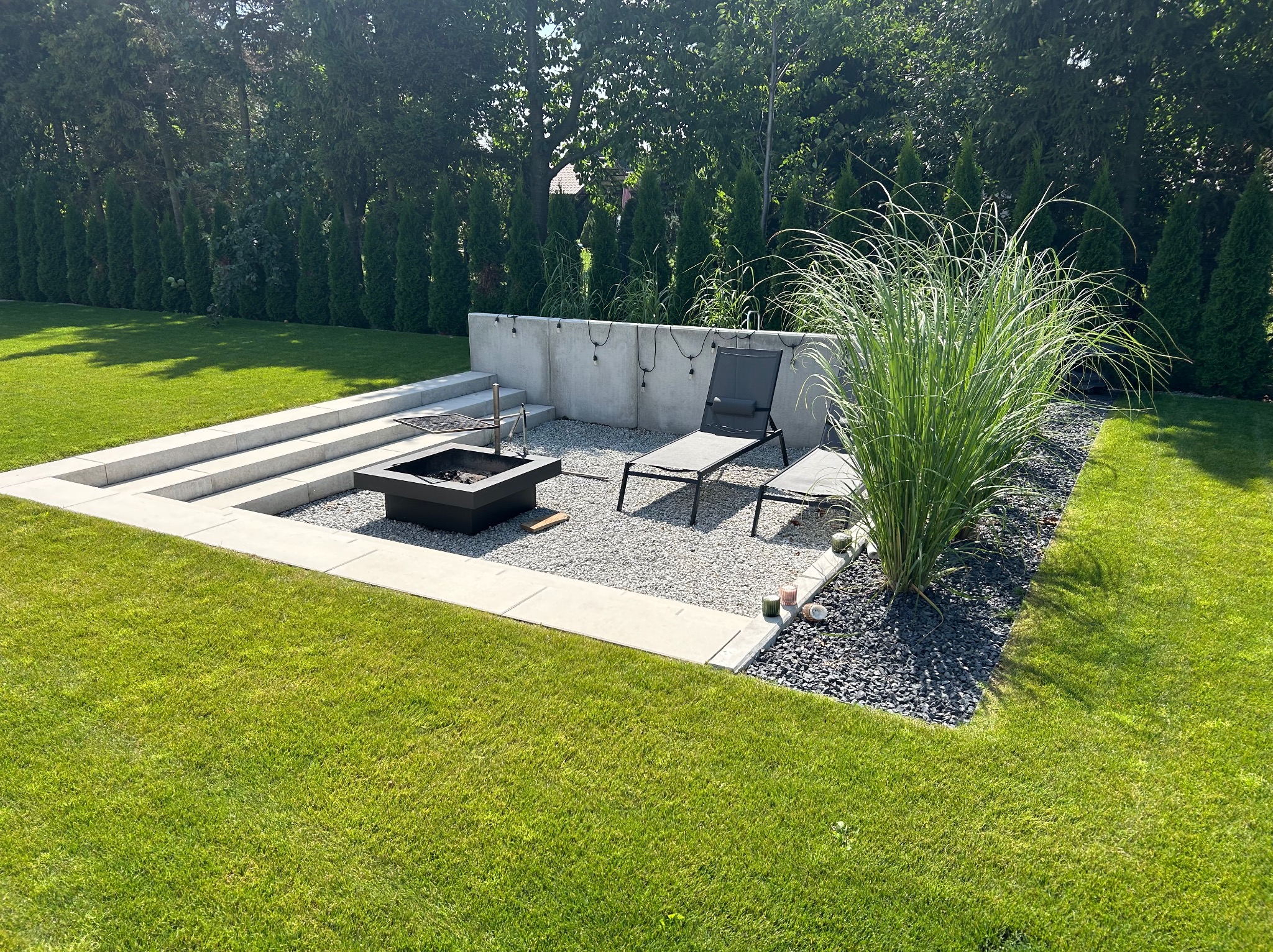 Strefa relaksu w ogrodzie w Ligocie: betonowe schody, palenisko, leżak, ozdobne trawy i kamienie. Nowoczesny design, idealne miejsce na wypoczynek.