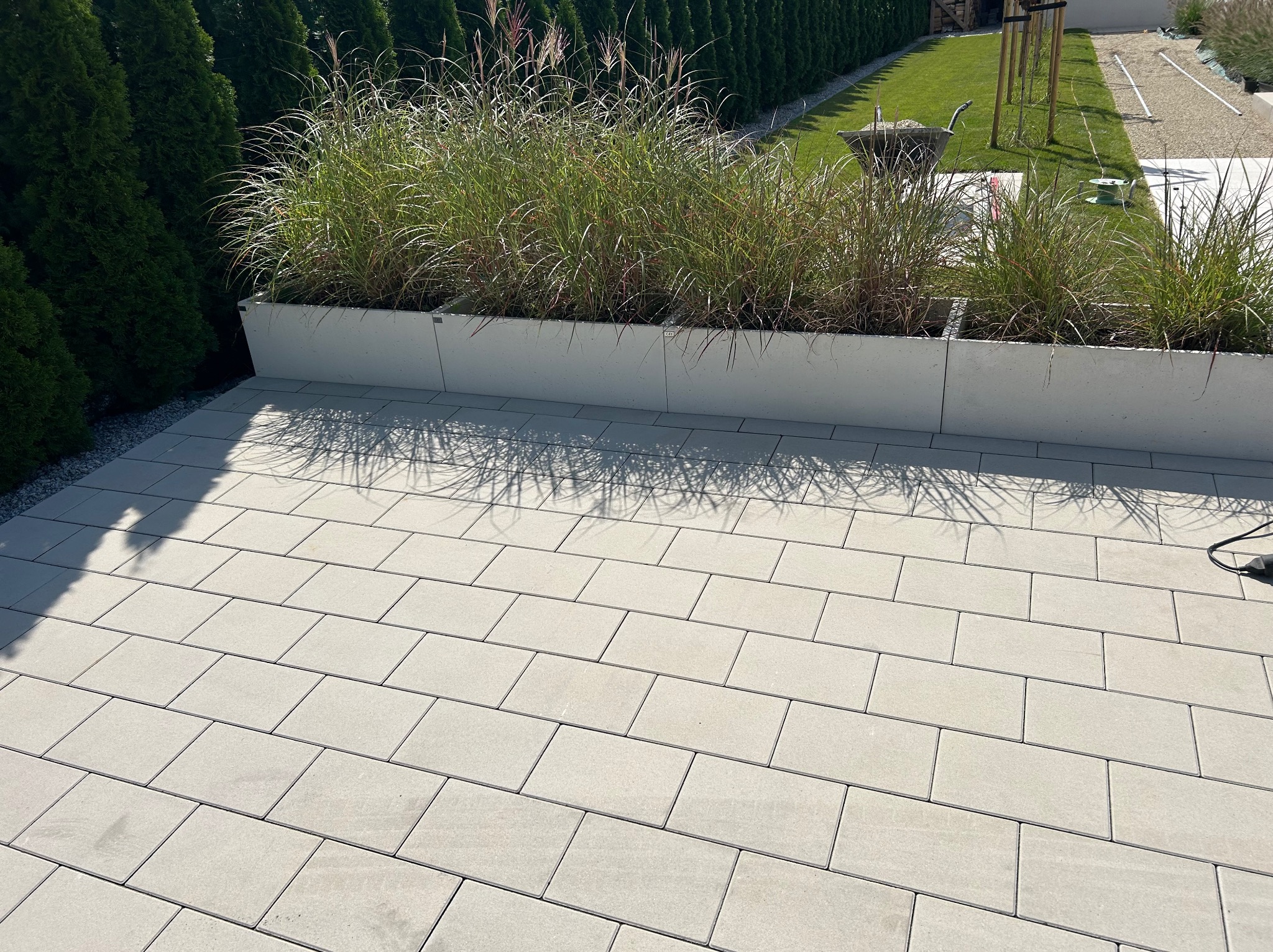 Jasnoszara kostka brukowa ułożona na tarasie, z donicami z trawami ozdobnymi w tle i zielonym żywopłotem. Widoczny fragment trawnika i przygotowania do dalszych prac.