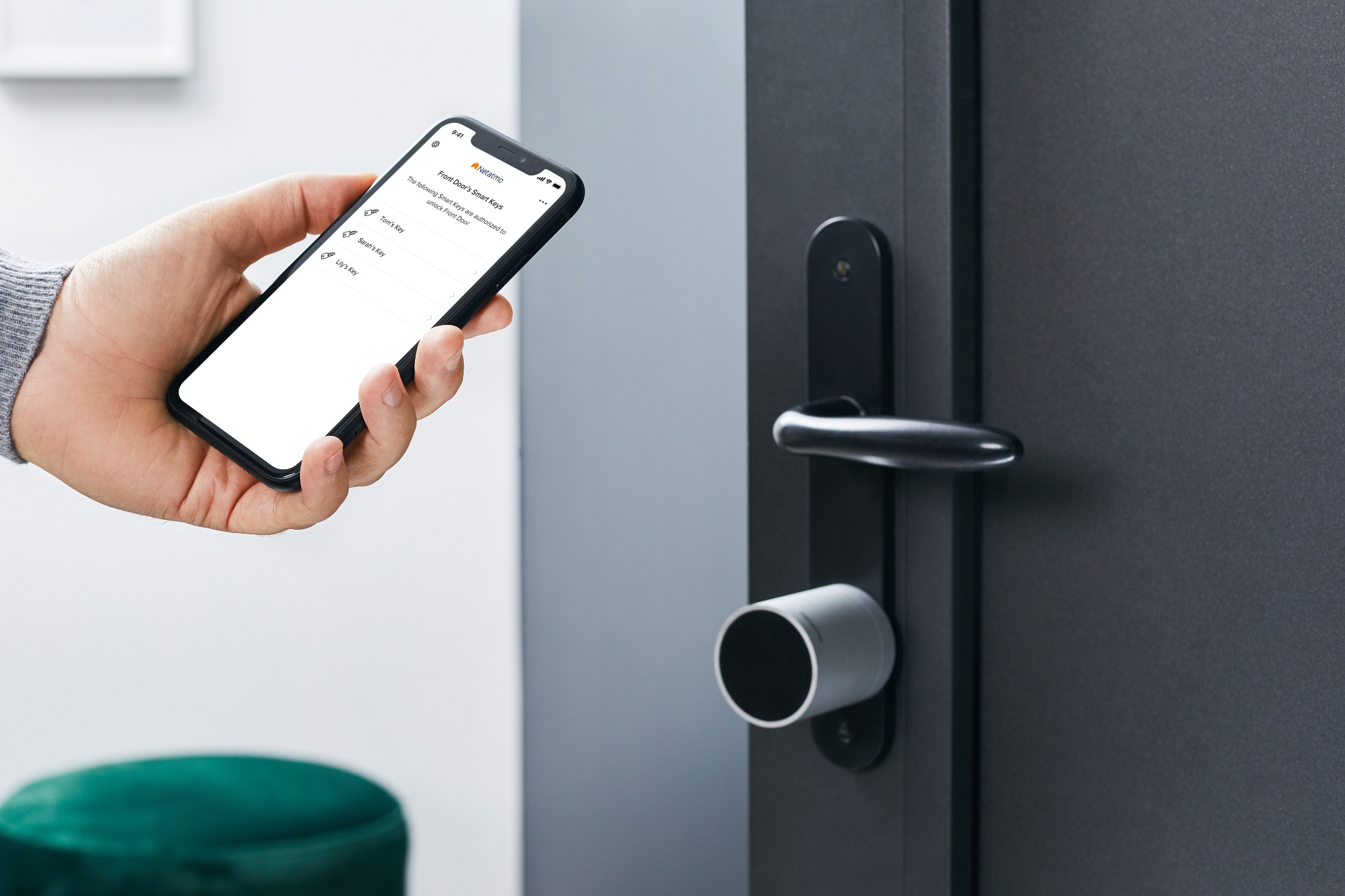 Dłoń trzymająca smartfon z aplikacją do zarządzania inteligentnym zamkiem do drzwi, widoczny nowoczesny zamek elektroniczny na ciemnych drzwiach.