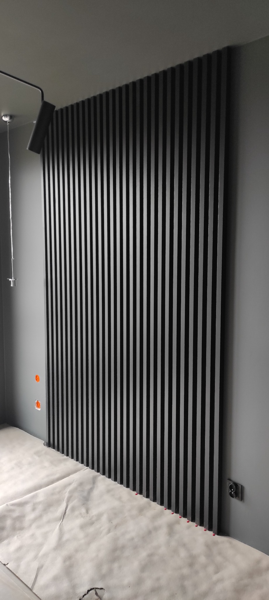 Ściana z czarnych, pionowych lameli dekoracyjnych w szarym wnętrzu, zabezpieczona podłoga papierem malarskim, widoczne gniazdko elektryczne i otwory na puszki elektryczne.