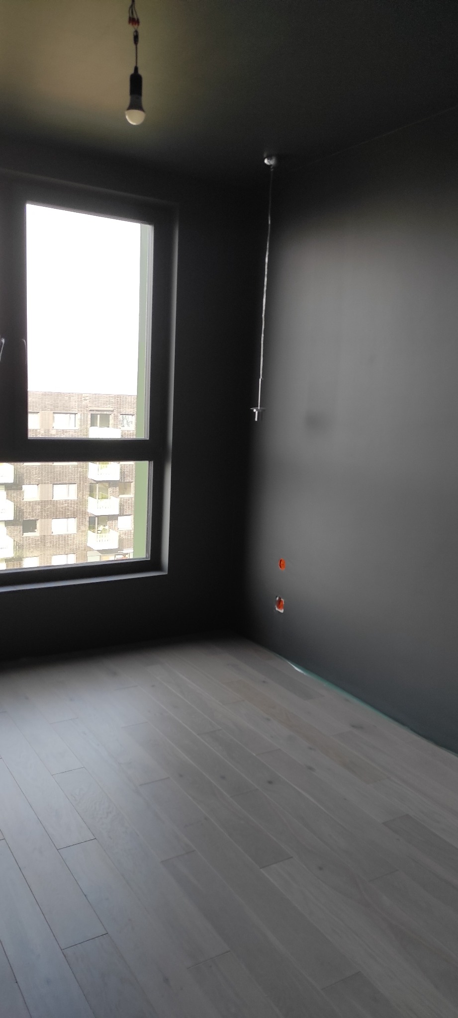 Wnętrze pomalowanego na ciemno pomieszczenia z jasną drewnianą podłogą, widoczne gniazdka elektryczne i okno z widokiem na budynek