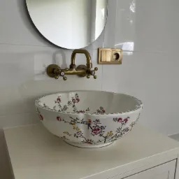 Umywalka ceramiczna w kwiaty i ptaki, bateria retro w kolorze starego złota, owalne lustro bez ramy, białe płytki i złote włączniki w łazience.