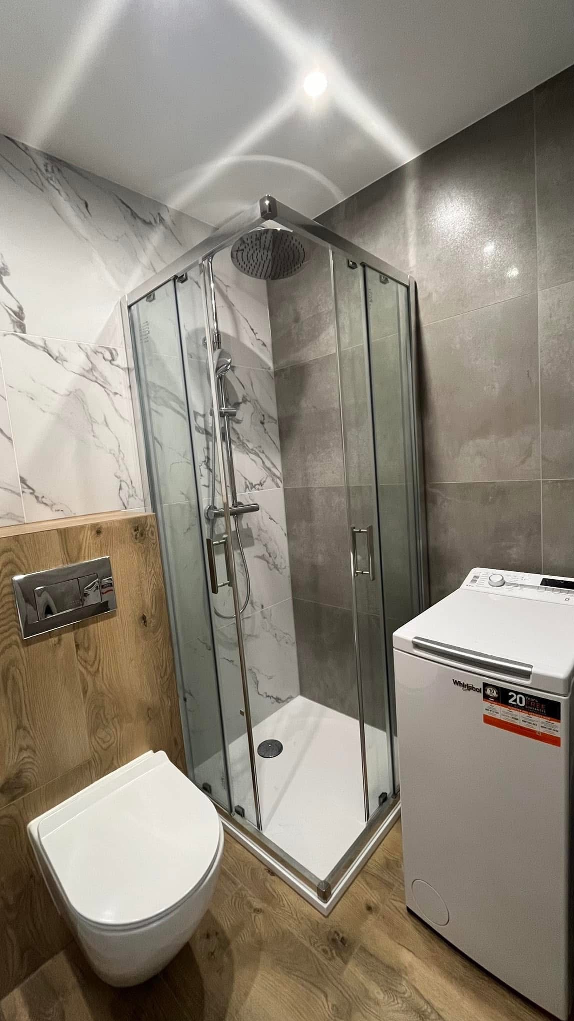 Nowoczesna łazienka z kabiną prysznicową narożną, wiszącą toaletą z drewnianym panelem, pralką Whirlpool i szarymi płytkami imitującymi marmur.