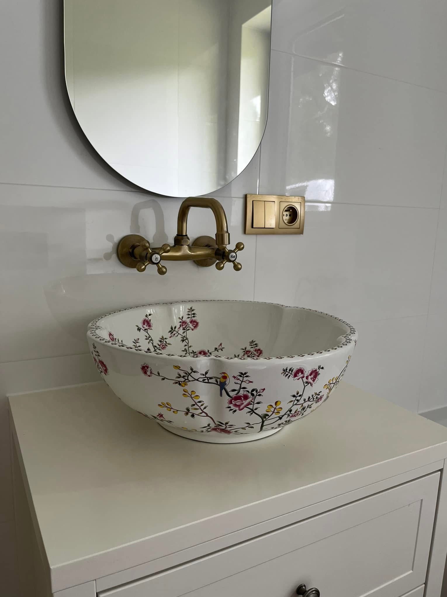 Umywalka ceramiczna w kwiaty i ptaki, bateria retro w kolorze starego złota, owalne lustro bez ramy, białe płytki i złote włączniki w łazience.