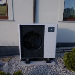 Zewnętrzna jednostka urządzenia grzewczego Mitsubishi Zubadan R32, umieszczona na betonowej podstawie z drenażem, otoczona białym żwirem i niskimi krzewami iglastymi przy ścianie budynku z dwoma...