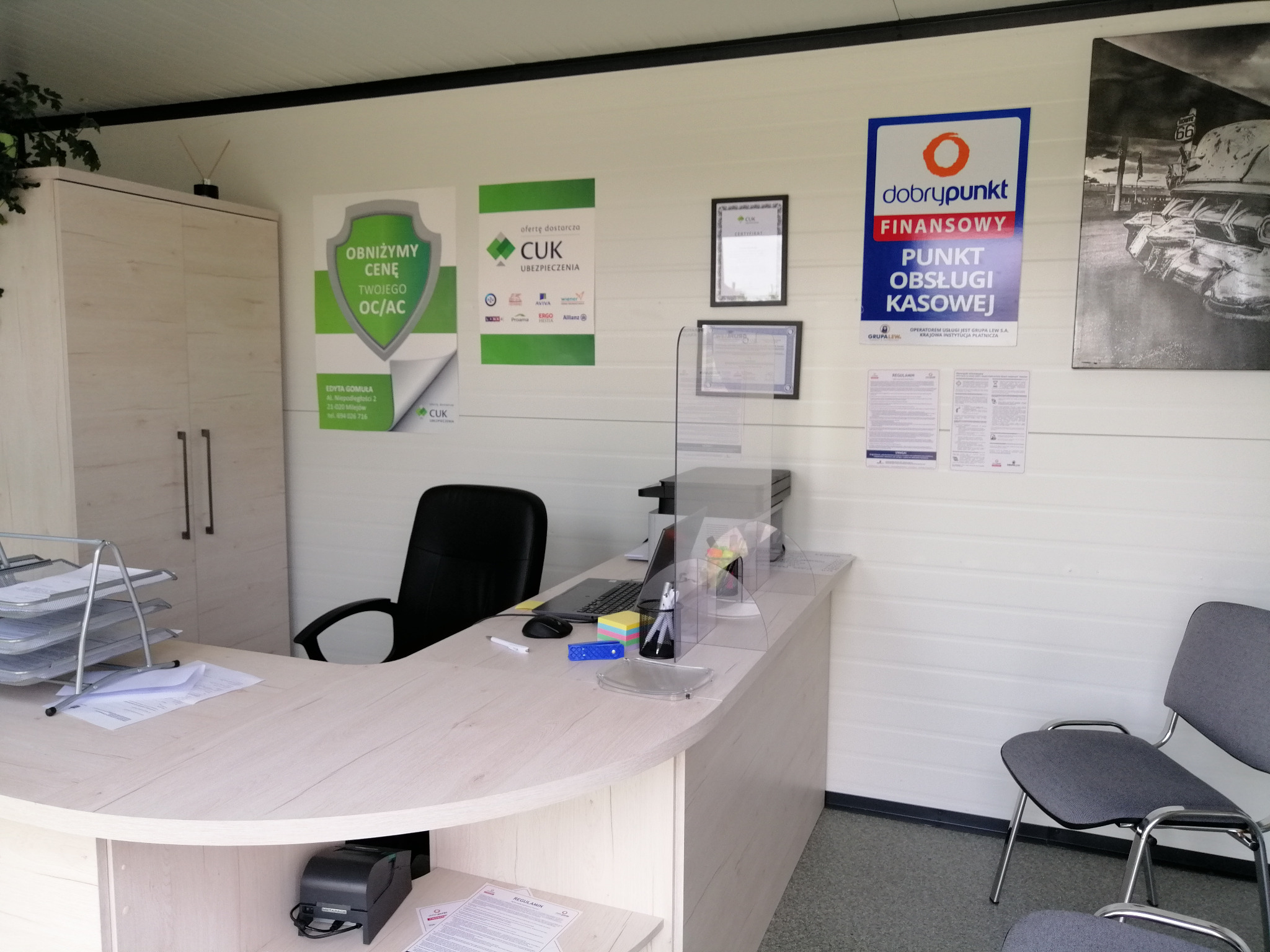 Wnętrze biura ubezpieczeniowego z biurkiem, krzesłami dla klientów i plakatami reklamowymi CUK i Dobry Punkt Finansowy na ścianie.