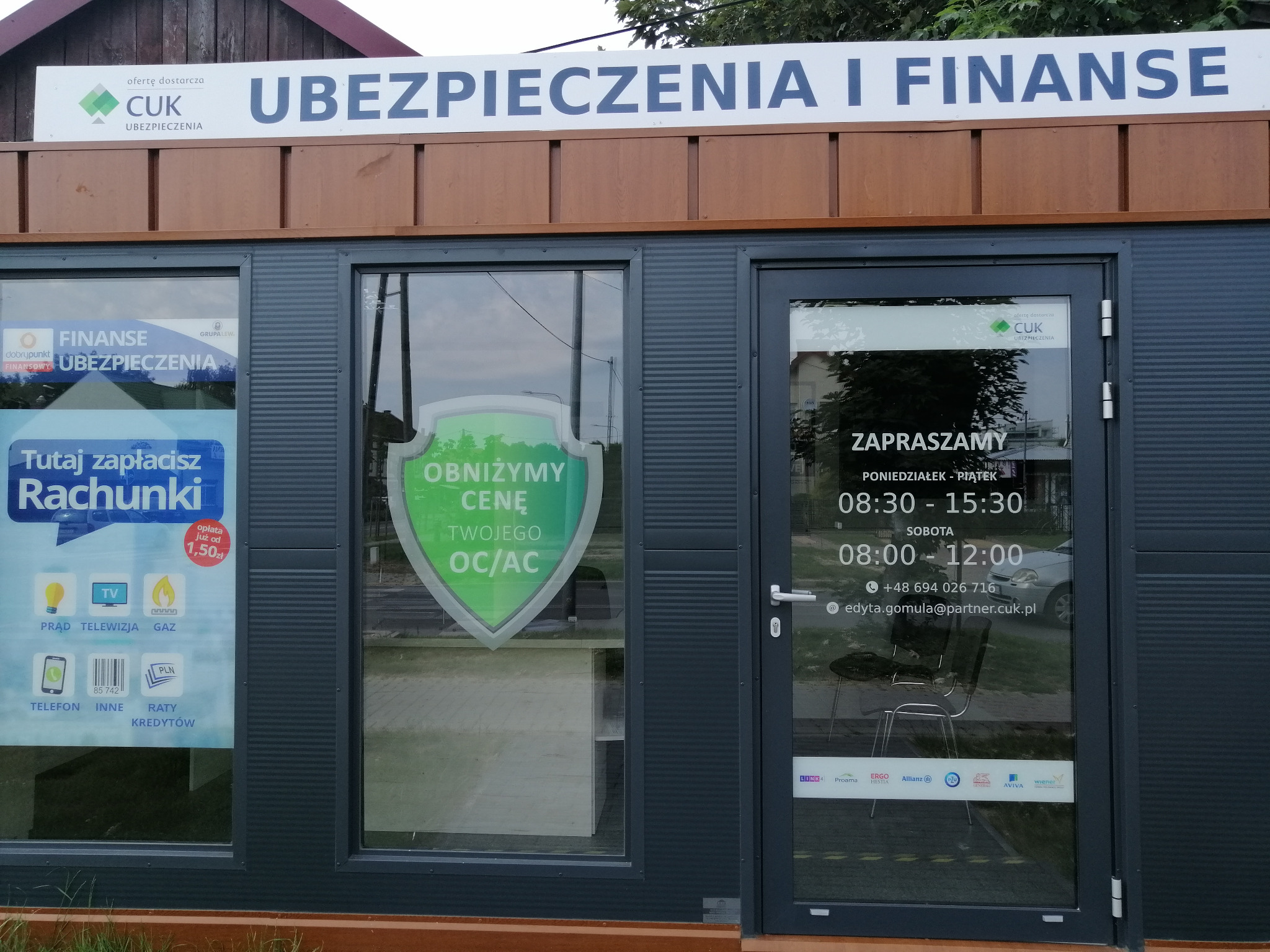 Biuro CUK Ubezpieczenia z napisem 'Ubezpieczenia i Finanse' nad wejściem. Na oknie naklejka informująca o możliwości zapłaty rachunków oraz obniżeniu ceny OC/AC. Na szybie godziny otwarcia...