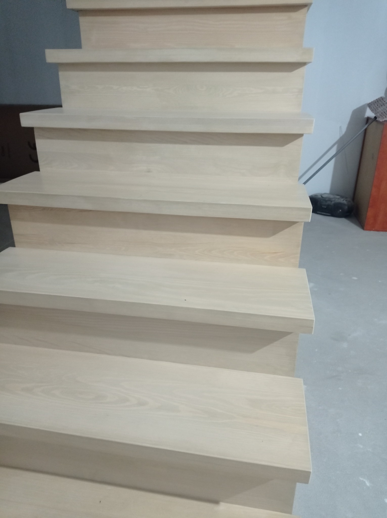 Jasne, drewniane schody z widocznym usłojeniem, ujęcie z dołu, ukazujące stopnie i krawędzie, w tle fragment ściany i podłogi.